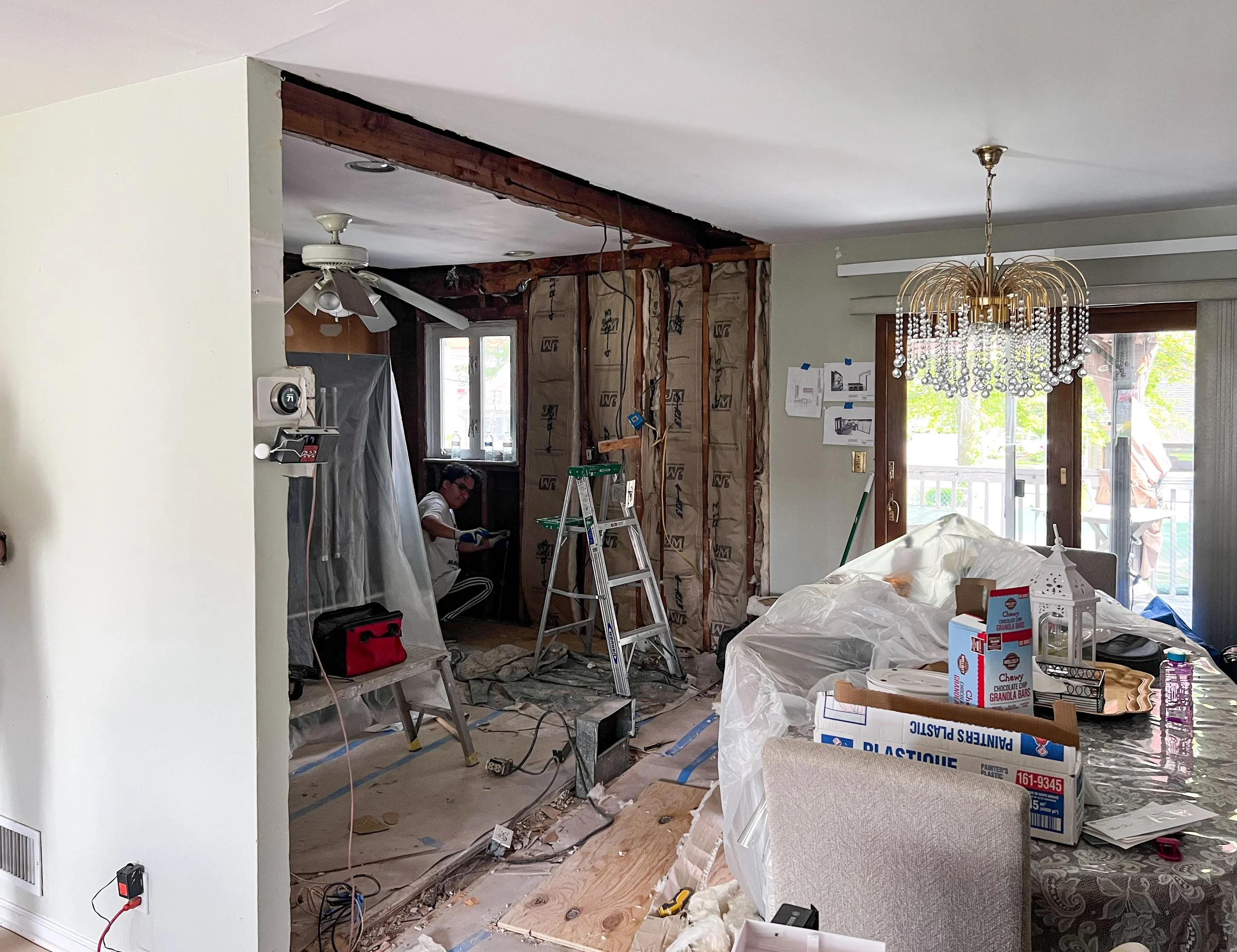 Interior of a home undergoing renovation, with exposed wall insulation, a worker sitting on the floor, a ladder, and construction tools. The dining area is cluttered with plastic-covered furniture, household items, and renovation materials.