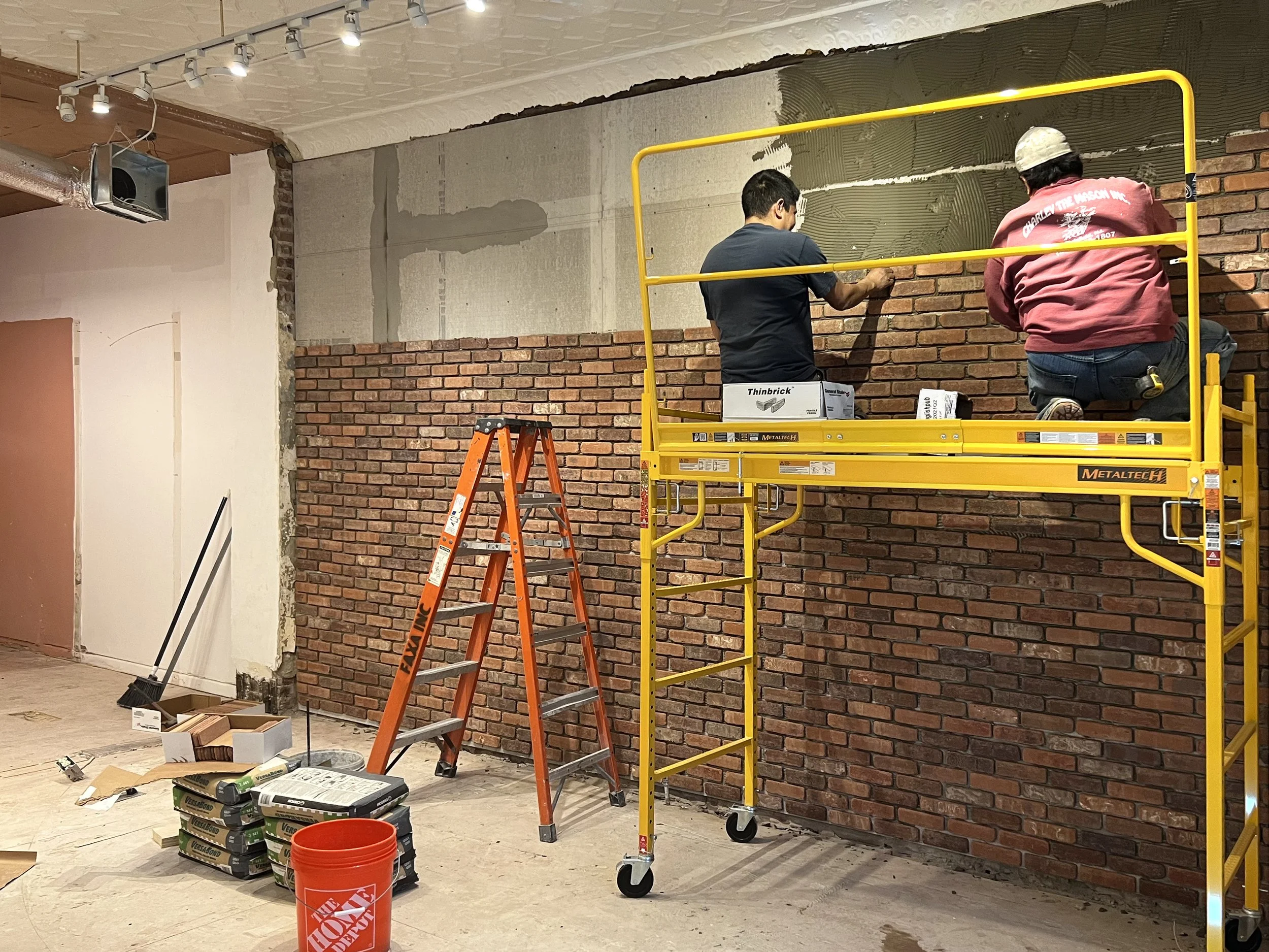 Two workers on a yellow scaffold installing brick veneer on an interior wall. Construction tools and materials are on the floor nearby, including a ladder, a broom, buckets, and bags of supplies.
