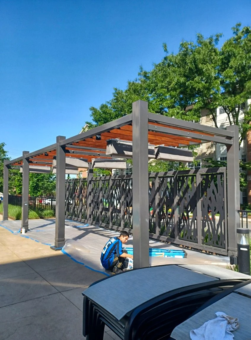 A person in a blue jacket sitting on the sidewalk near a modern metal and wood structure, possibly a pergola with a decorative fence in the background, under a clear blue sky with green trees.