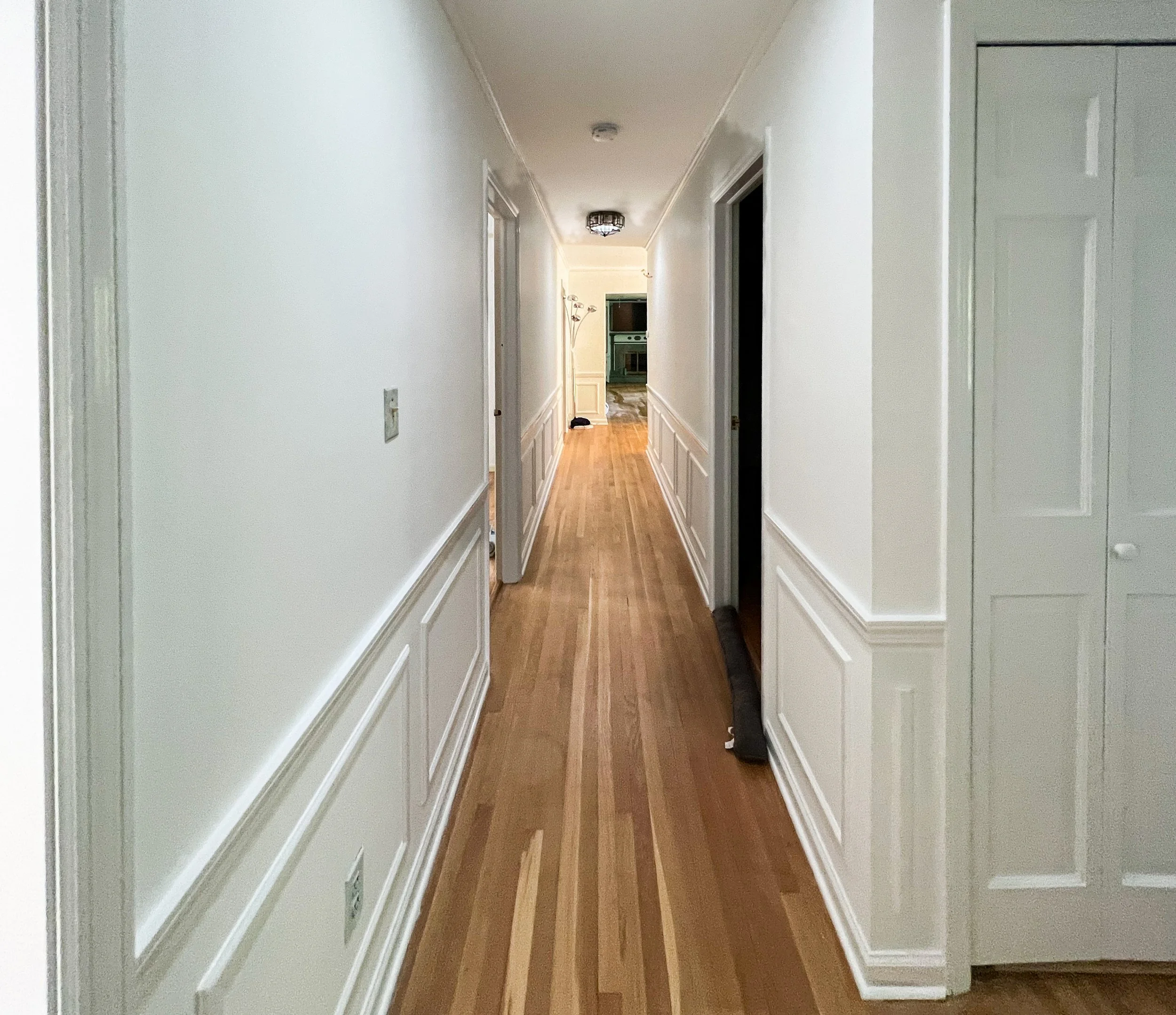 A hallway with white walls and wooden flooring, leading to a kitchen in the background.