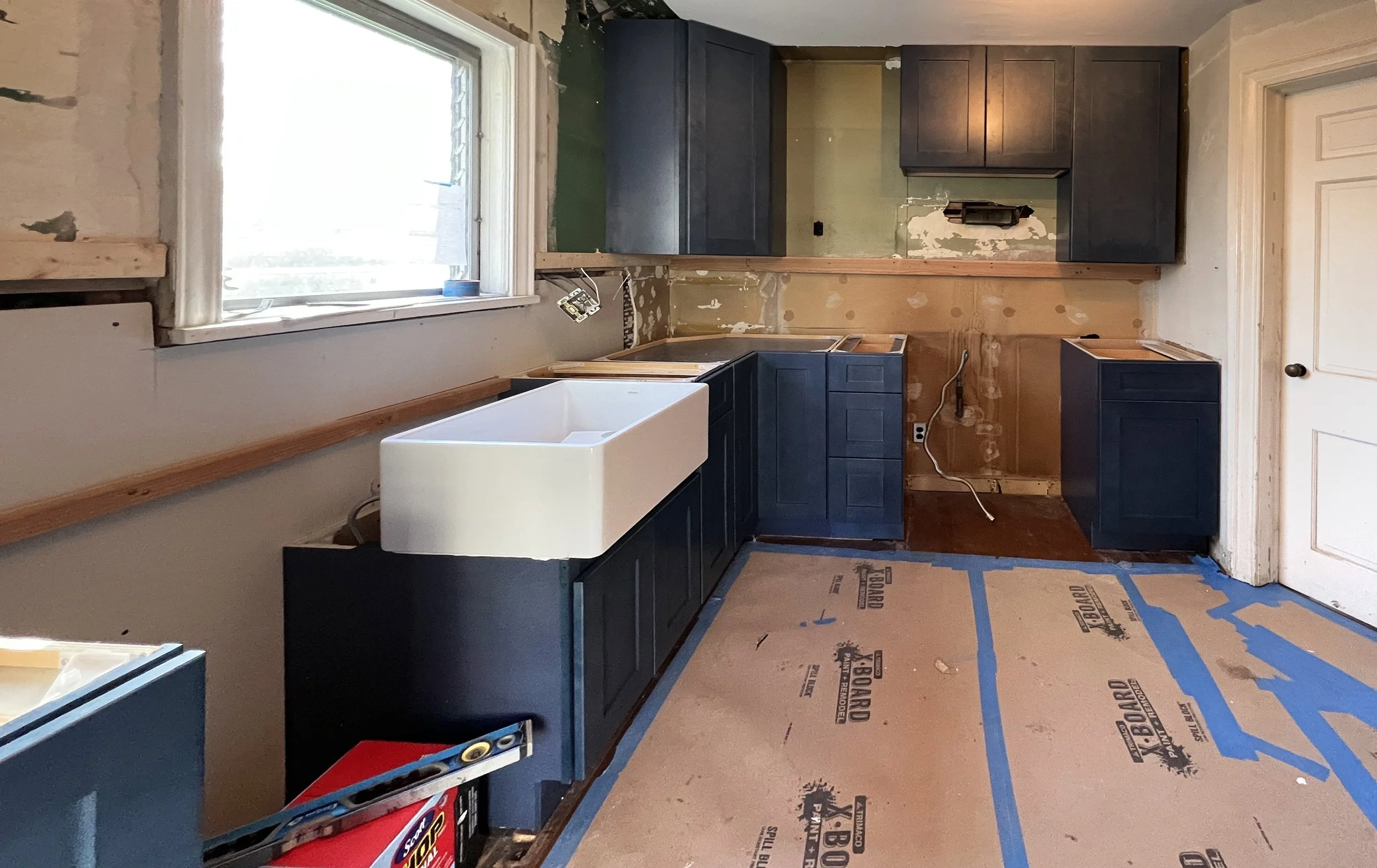 Kitchen under renovation with dark blue cabinets, a large white farmhouse sink, drywall prep, and construction materials on the floor.