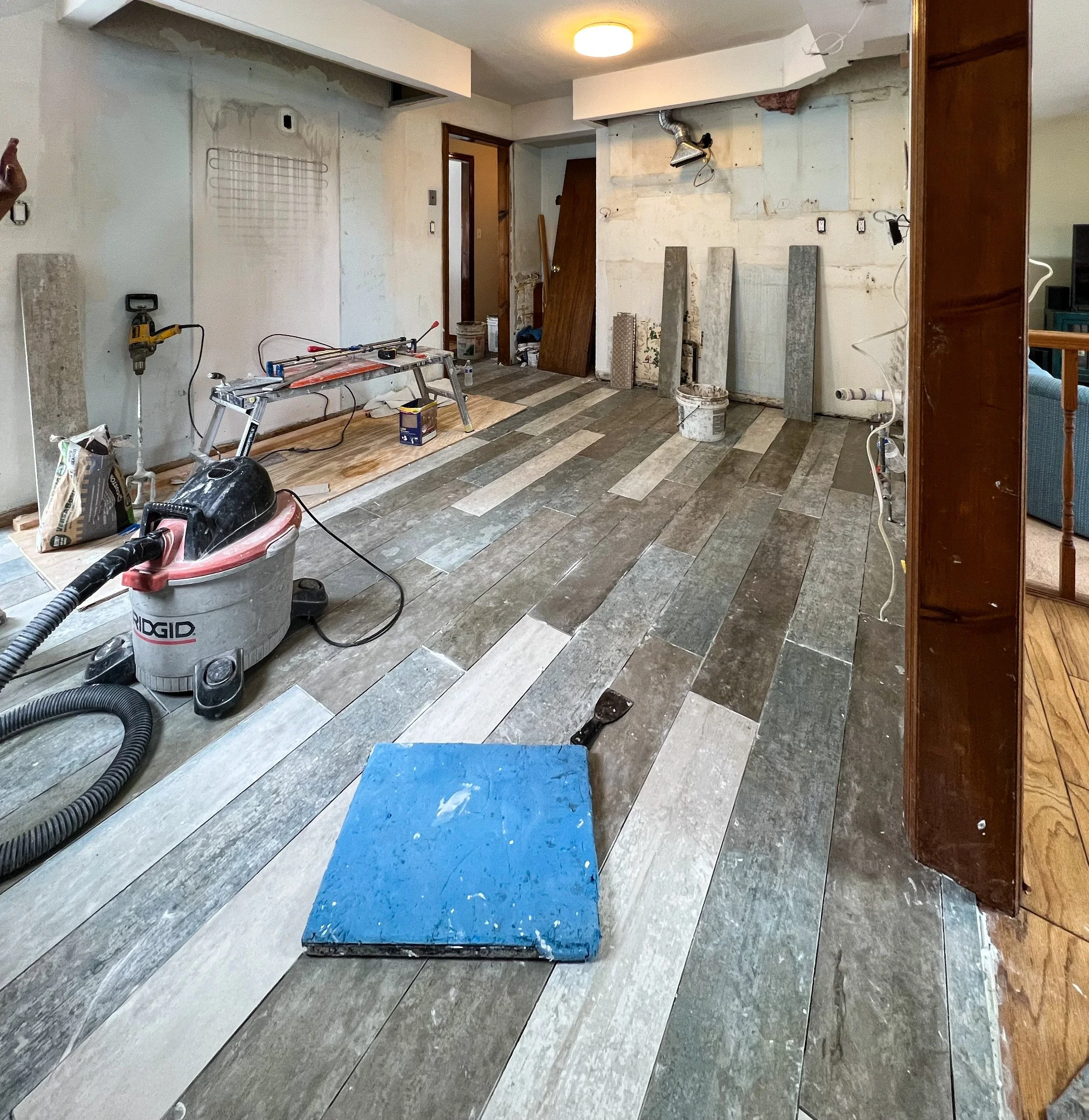 Room under renovation with partially installed wood-look tile flooring, construction tools, a shop vacuum, and construction materials scattered around.