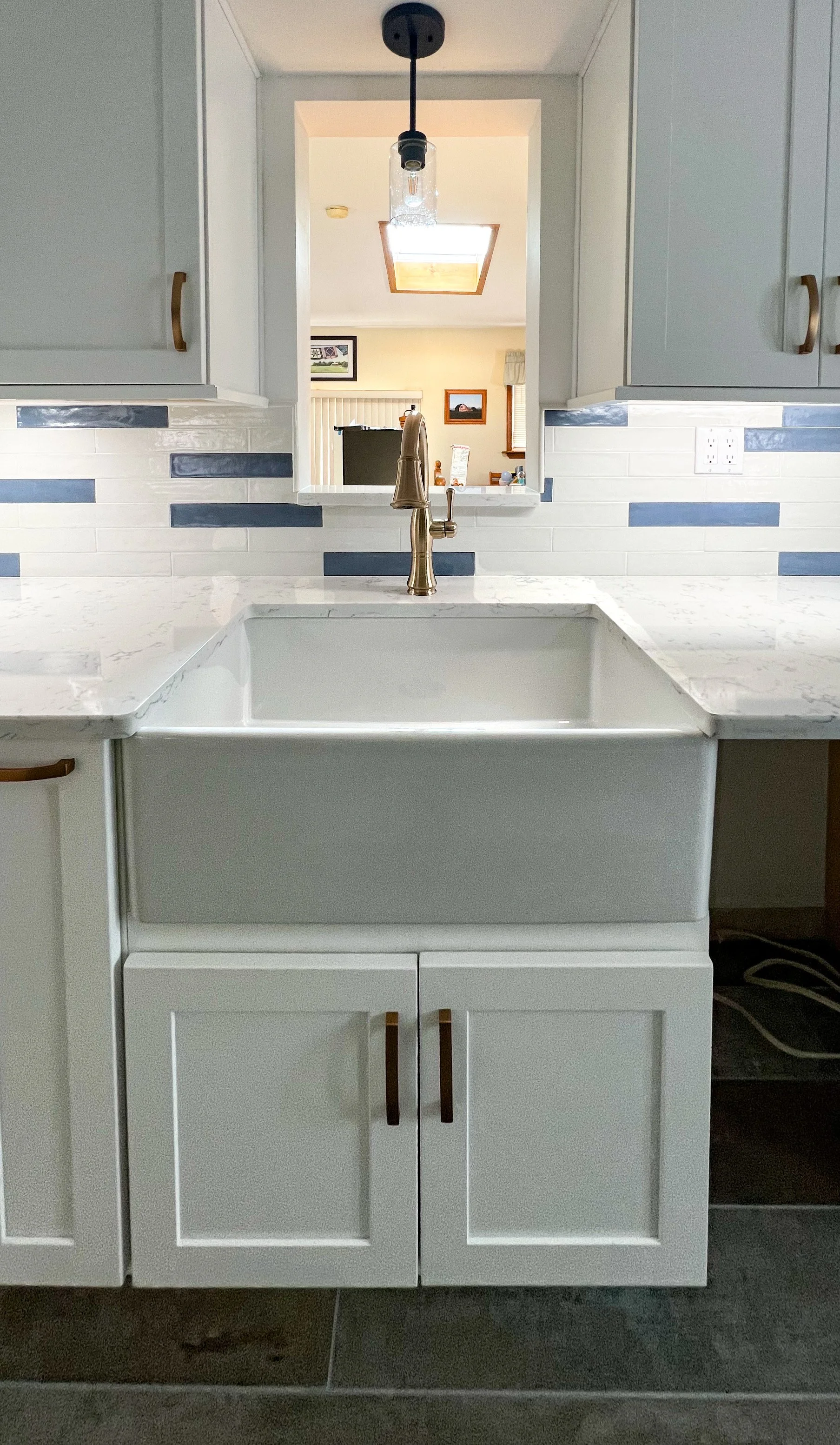 Kitchen sink with white cabinet and gold handles, marble countertop, and a windowed pass-through to another room.
