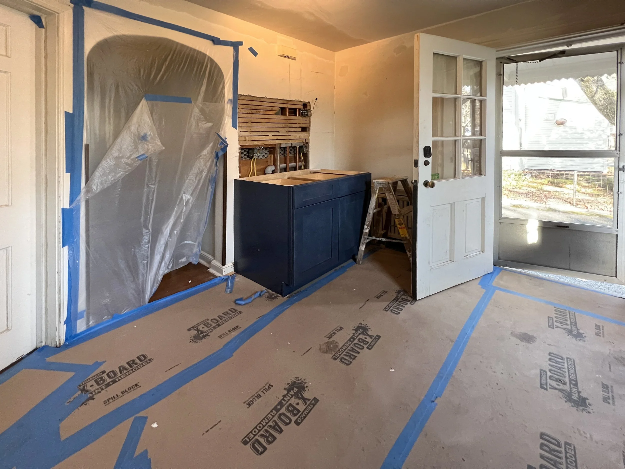 Interior of a room under renovation with construction tape on the floor, a partially installed kitchen cabinet, a ladder, a door, and plastic covering an opening.