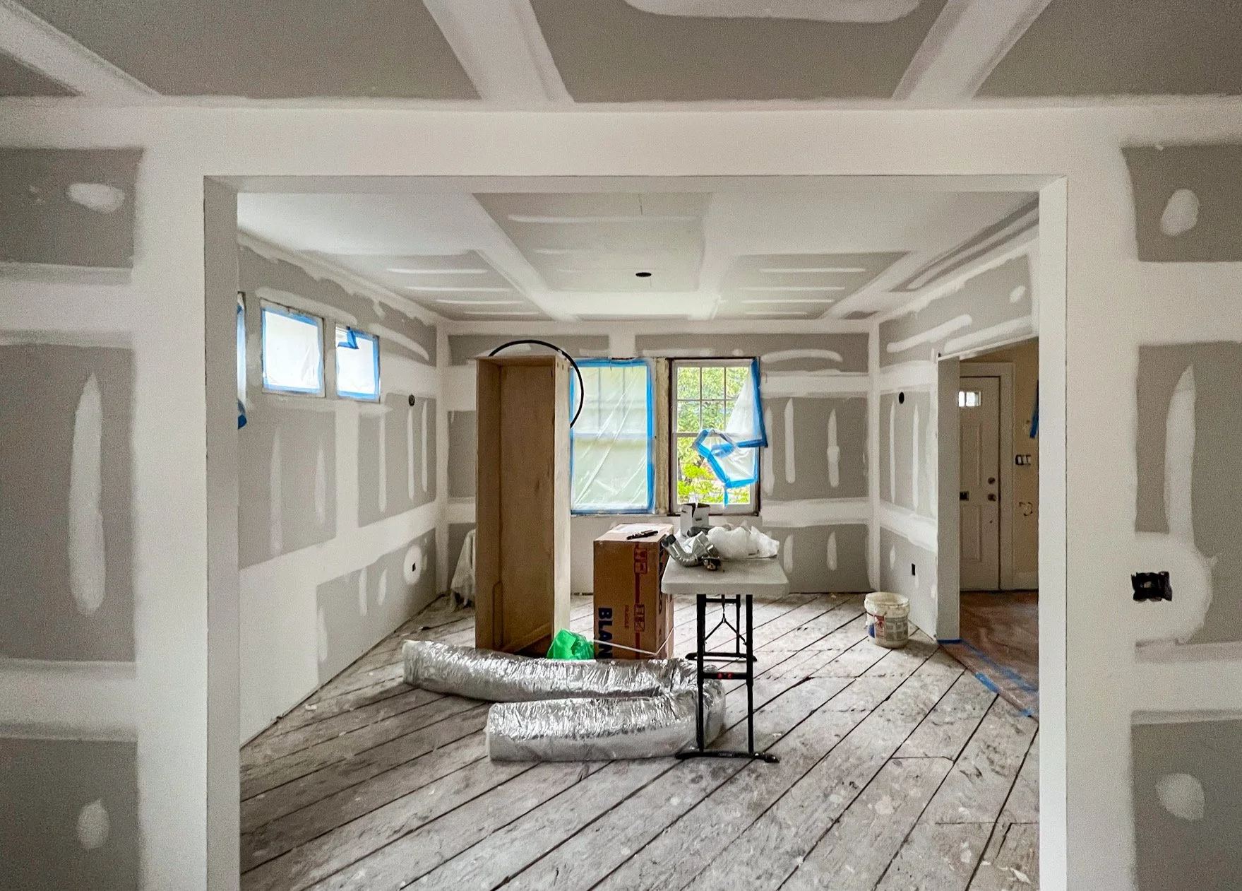 Interior of a house under construction with drywall walls, partially taped and mudded, and a wooden floor in progress. There are construction materials, tools, and windows with blue tape.