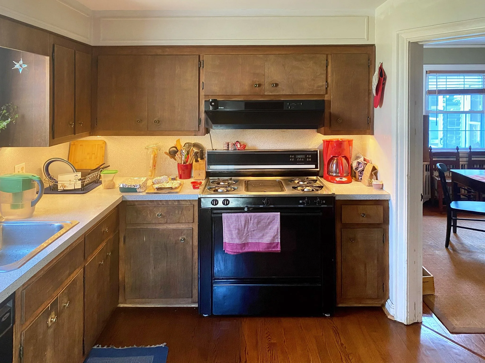 A kitchen with brown wooden cabinets, black stove, and various kitchen items on the countertops, including a red coffee maker and a green pitcher.