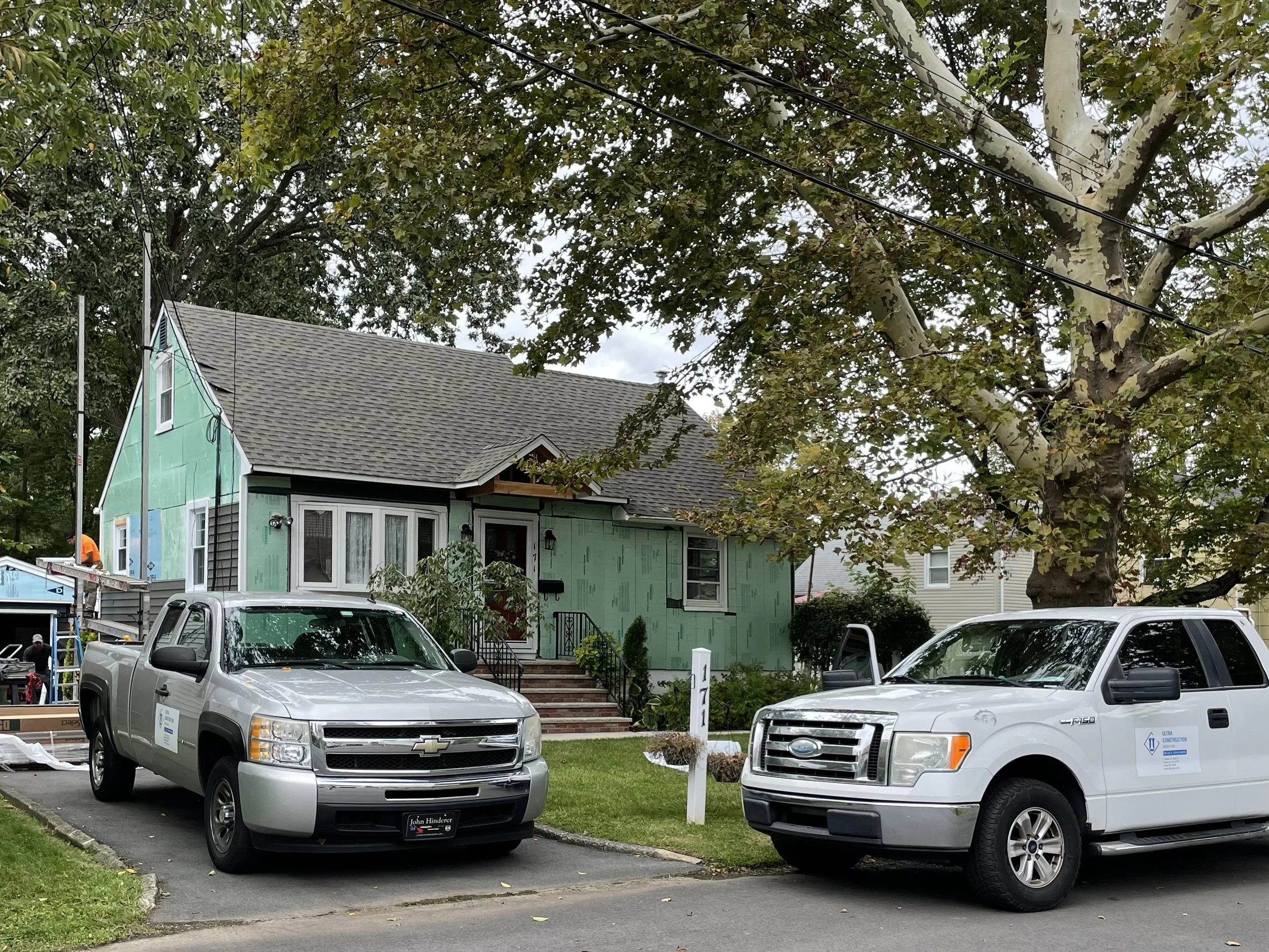 Two pickup trucks parked in front of a house under construction, with a large tree overhead and construction workers on the left side.