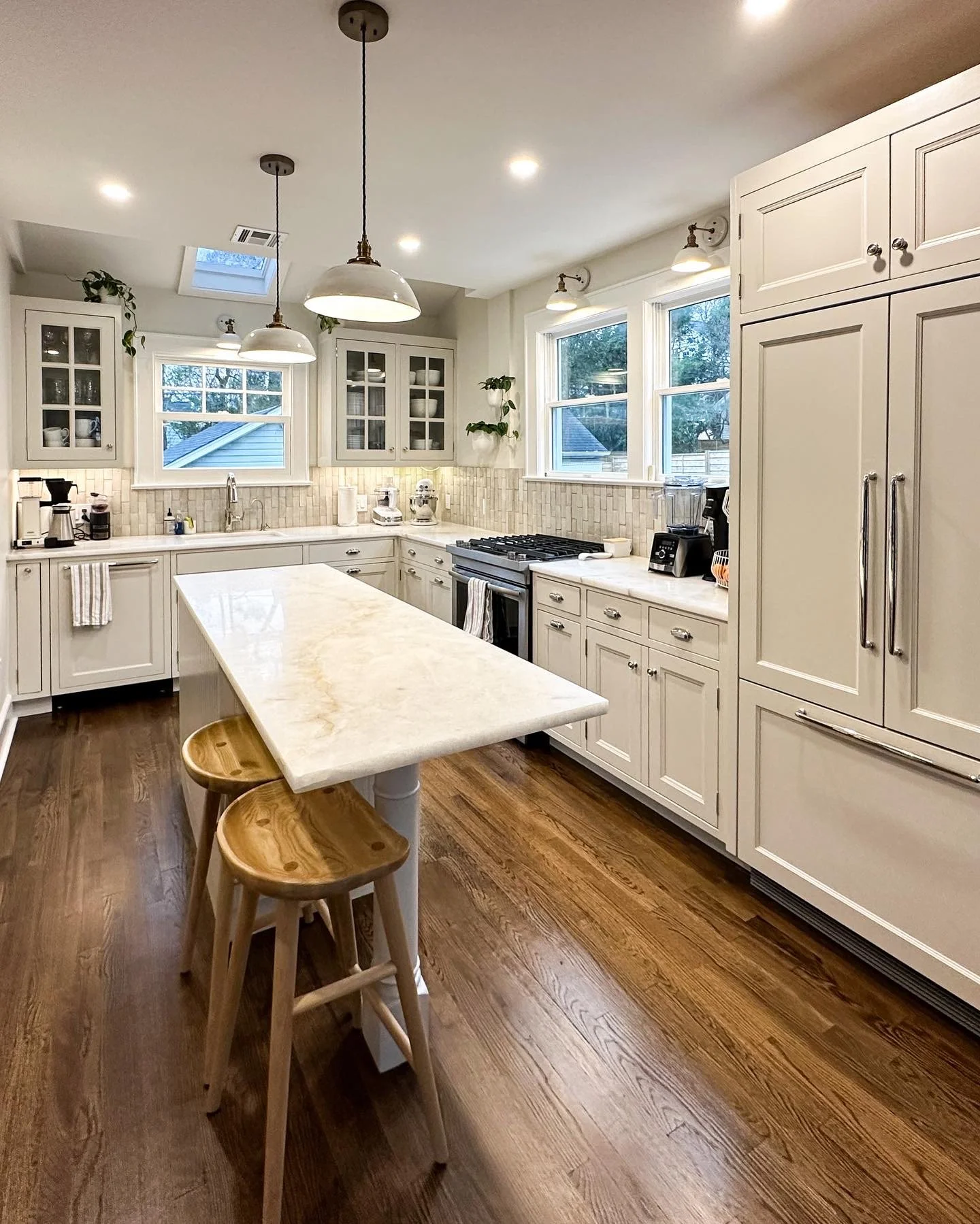 Bright kitchen with white cabinets, a kitchen island with a marble top, and three wooden bar stools. There are two windows above the countertop, a ceiling skylight, and recessed lighting. Small appliances like a coffee maker, blender, and stove are v