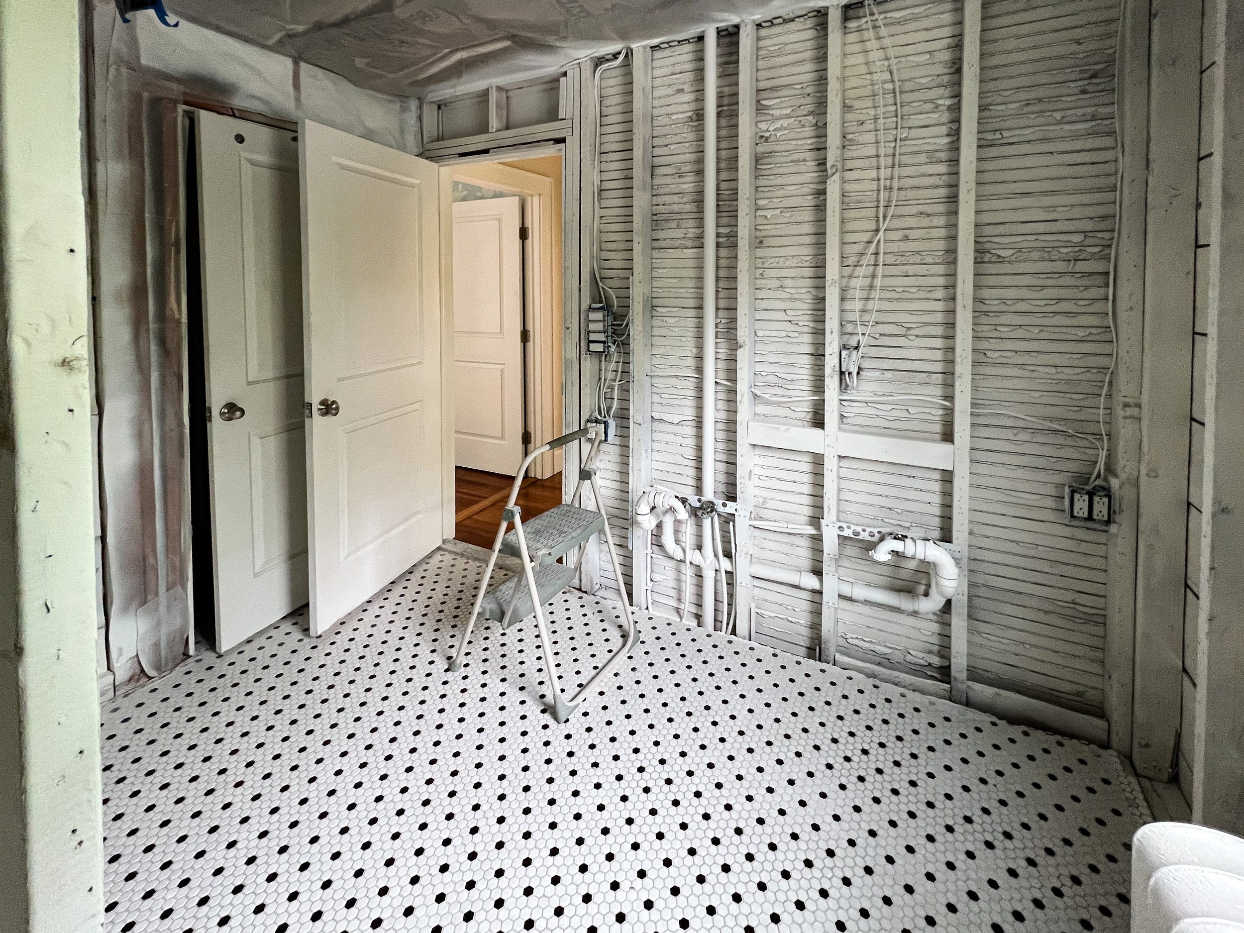 An unfinished room with exposed wall framing, plumbing pipes, a white door partially open, and a step stool on patterned black and white tiles.