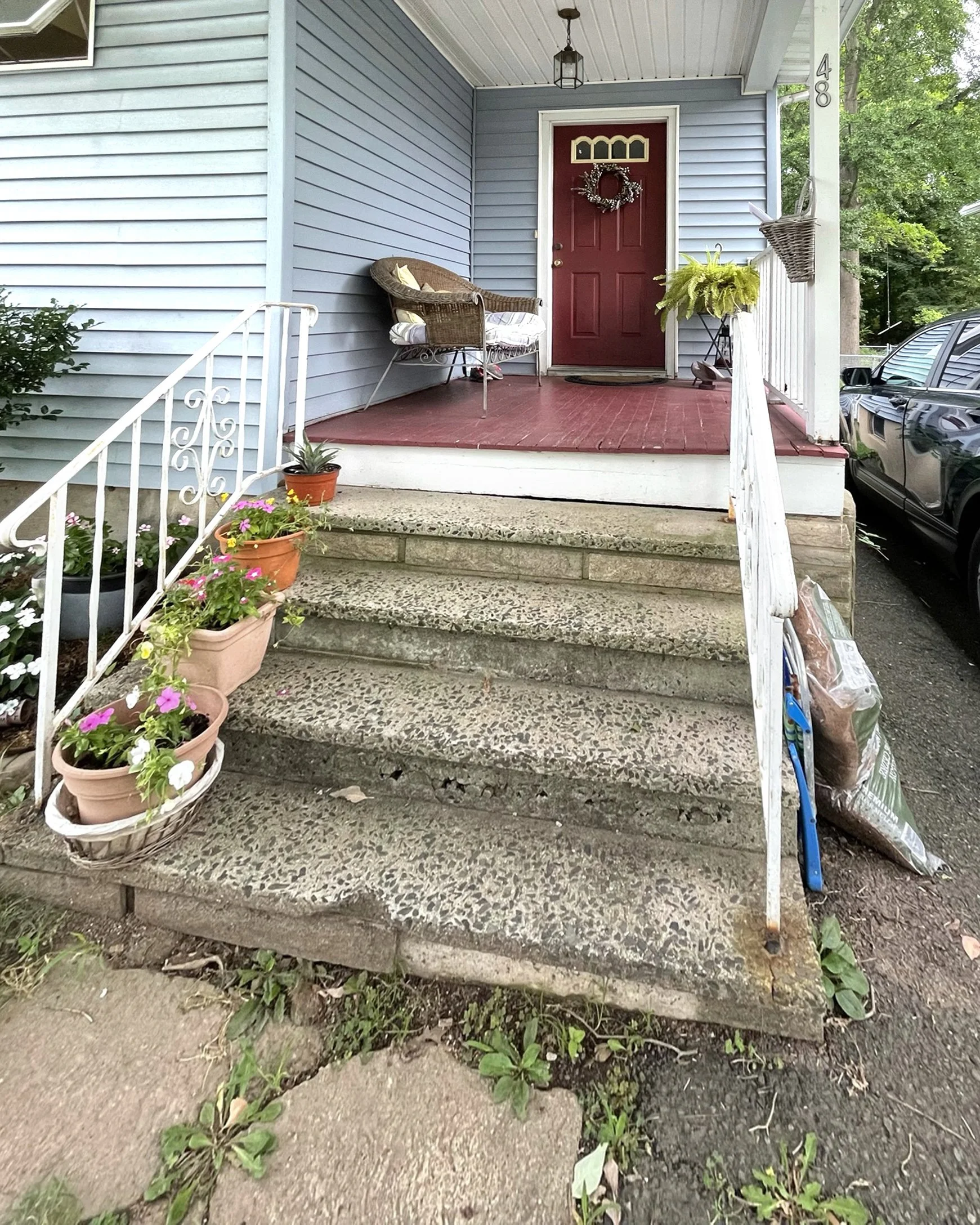 Front porch of a house with concrete steps leading up to a red deck and a red front door with a holiday wreath. There are potted flowers on the steps, a wicker chair on the porch, hanging baskets, and a porch light fixture.
