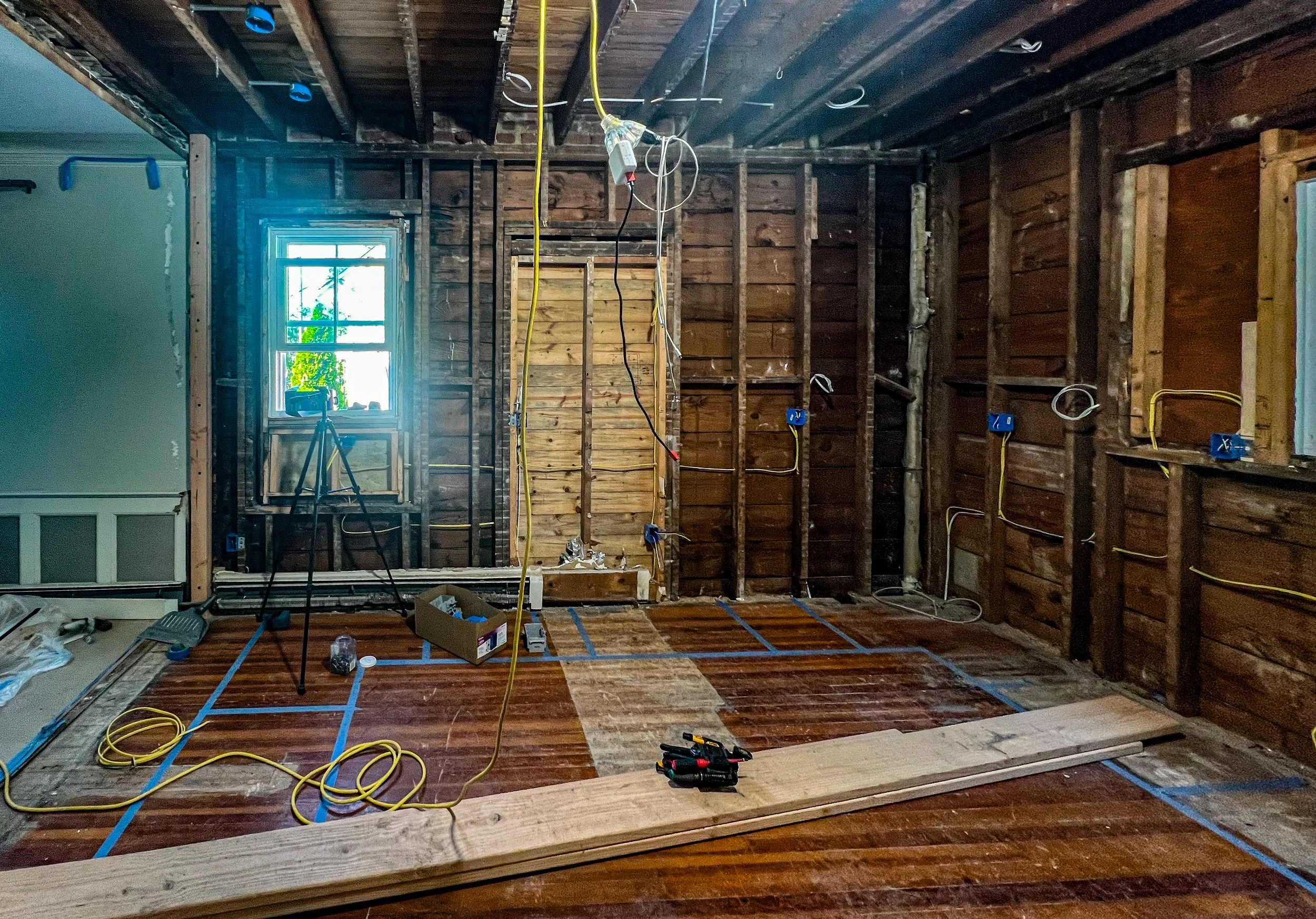 Room under renovation with exposed wooden studs and wiring on walls, a window letting in natural light, and partially removed flooring with tools and safety equipment visible.