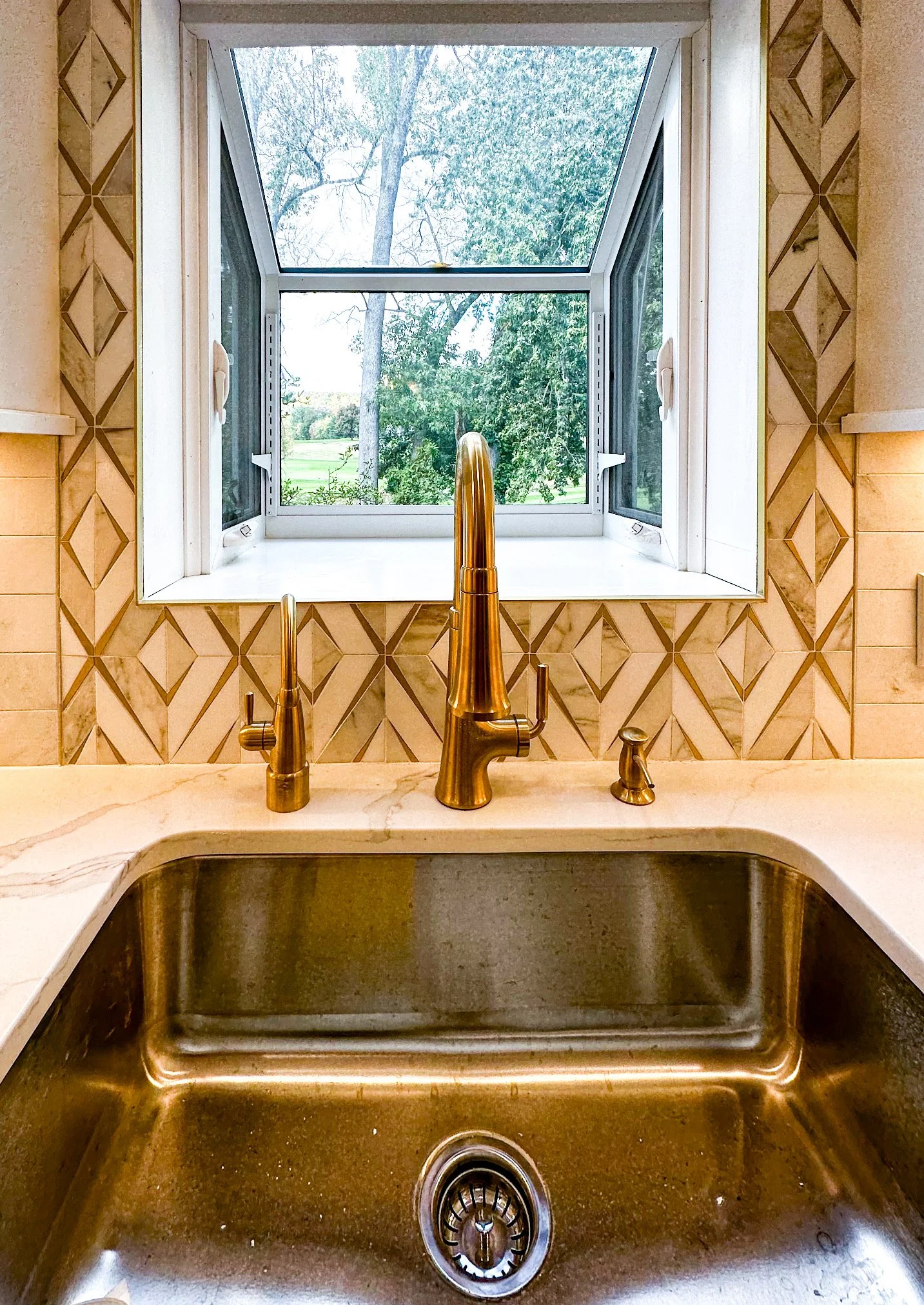 A kitchen sink with a brass faucet and a window looking outside to green trees.