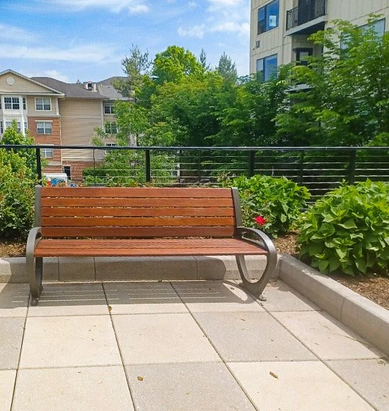 A wooden park bench with metal armrests on a tiled patio, surrounded by green bushes and trees, with residential buildings in the background.
