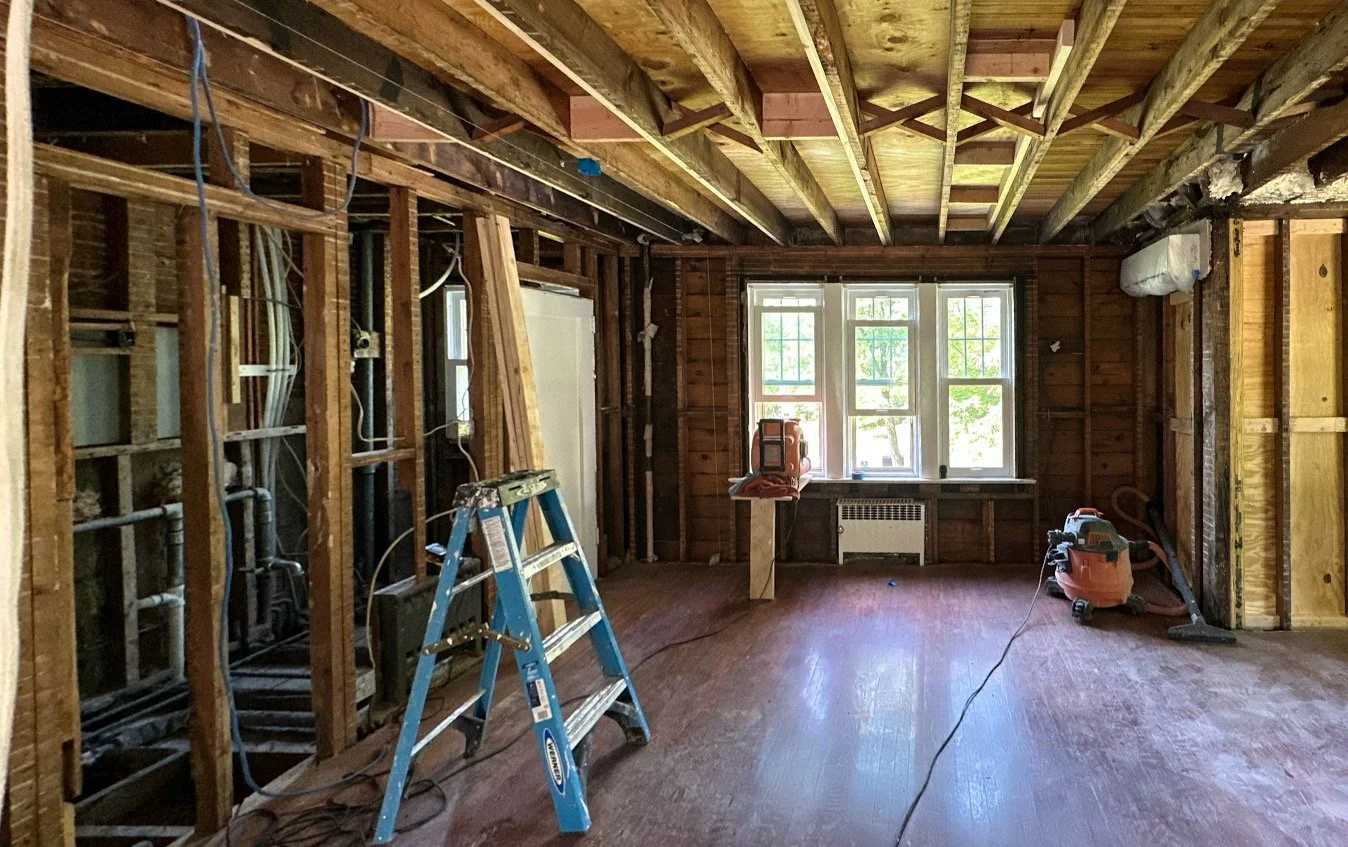 Interior of a house under renovation with exposed wooden studs, framing, and ceiling beams. A blue ladder is in the middle, and a vacuum cleaner and tools are on the floor. A window at the back lets in natural light, illuminating the unfinished space
