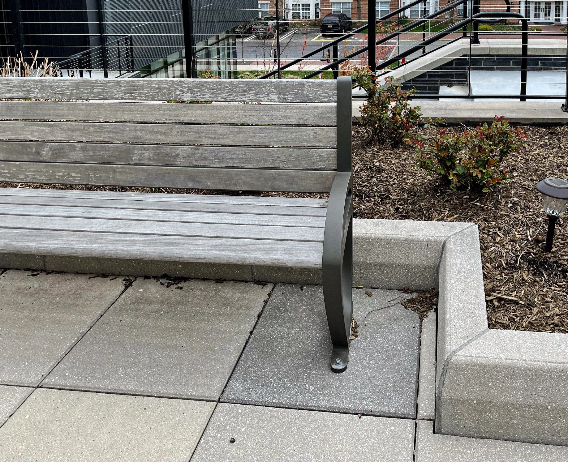 A wooden park bench with black metal supports on a concrete sidewalk, next to a small landscaped area with bushes and mulch.