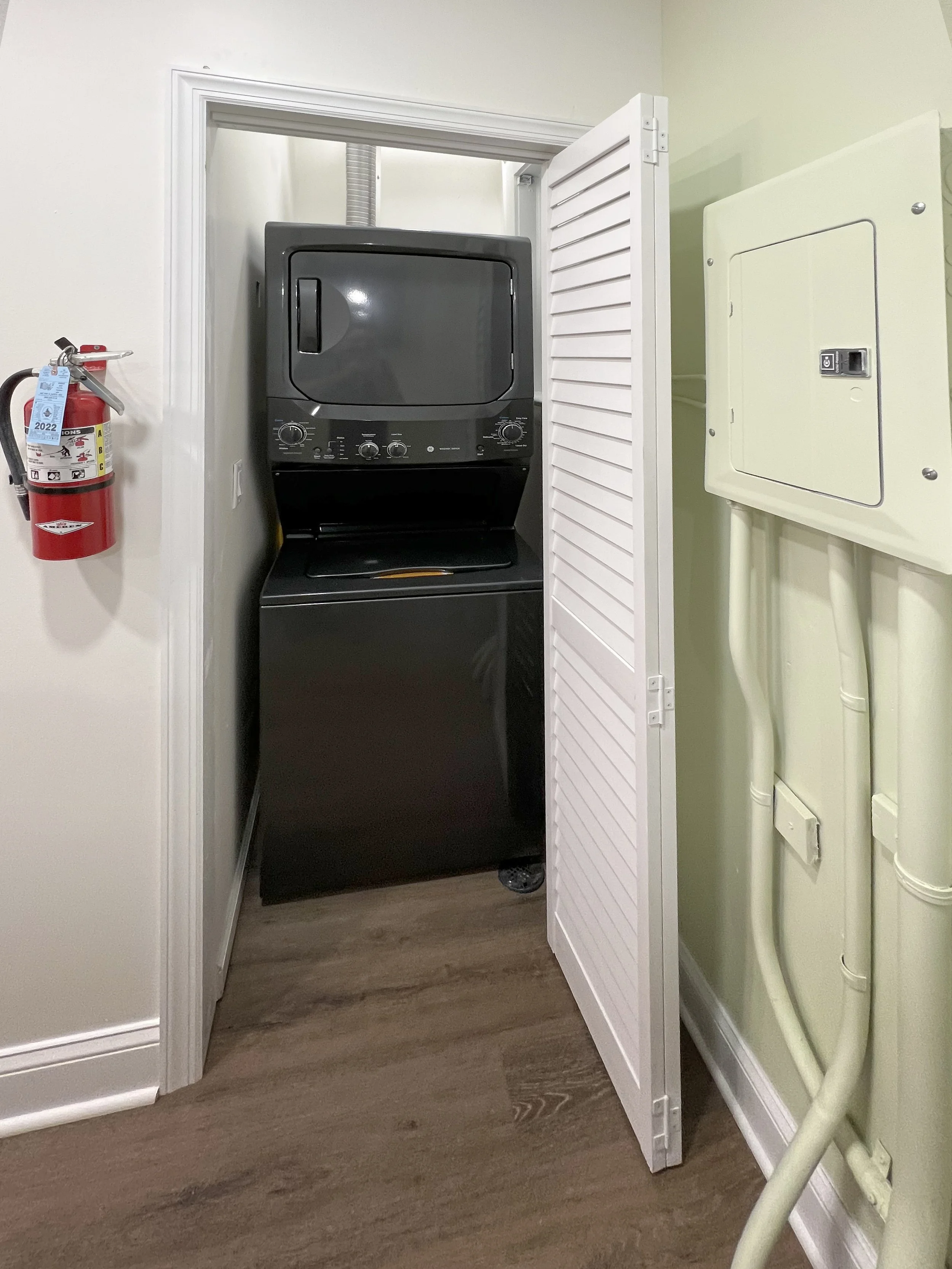 A laundry closet with a stacked black washer and dryer, a white louvered door partially open, a fire extinguisher mounted on the wall, and a utility panel on the wall.