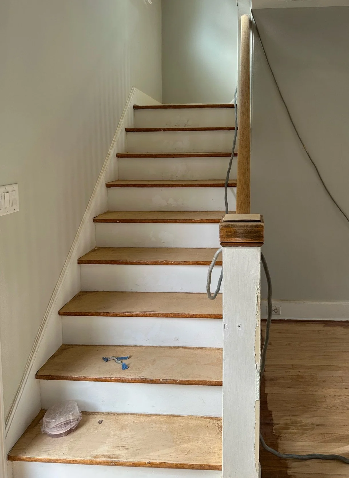 Unfinished staircase with wooden steps and a partially painted white riser, next to a wall with a light switch. Some construction materials and wires are visible.
