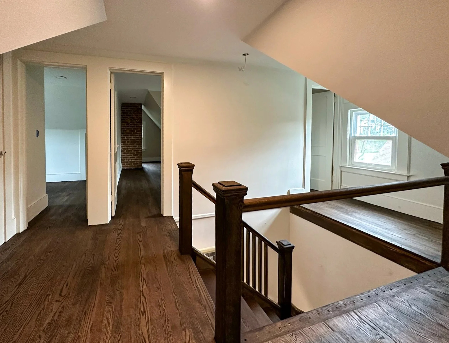 Upper floor of a house with wooden floors, stairs, windows, and doorways leading to different rooms, with a sloped ceiling and brick chimney visible in the background.