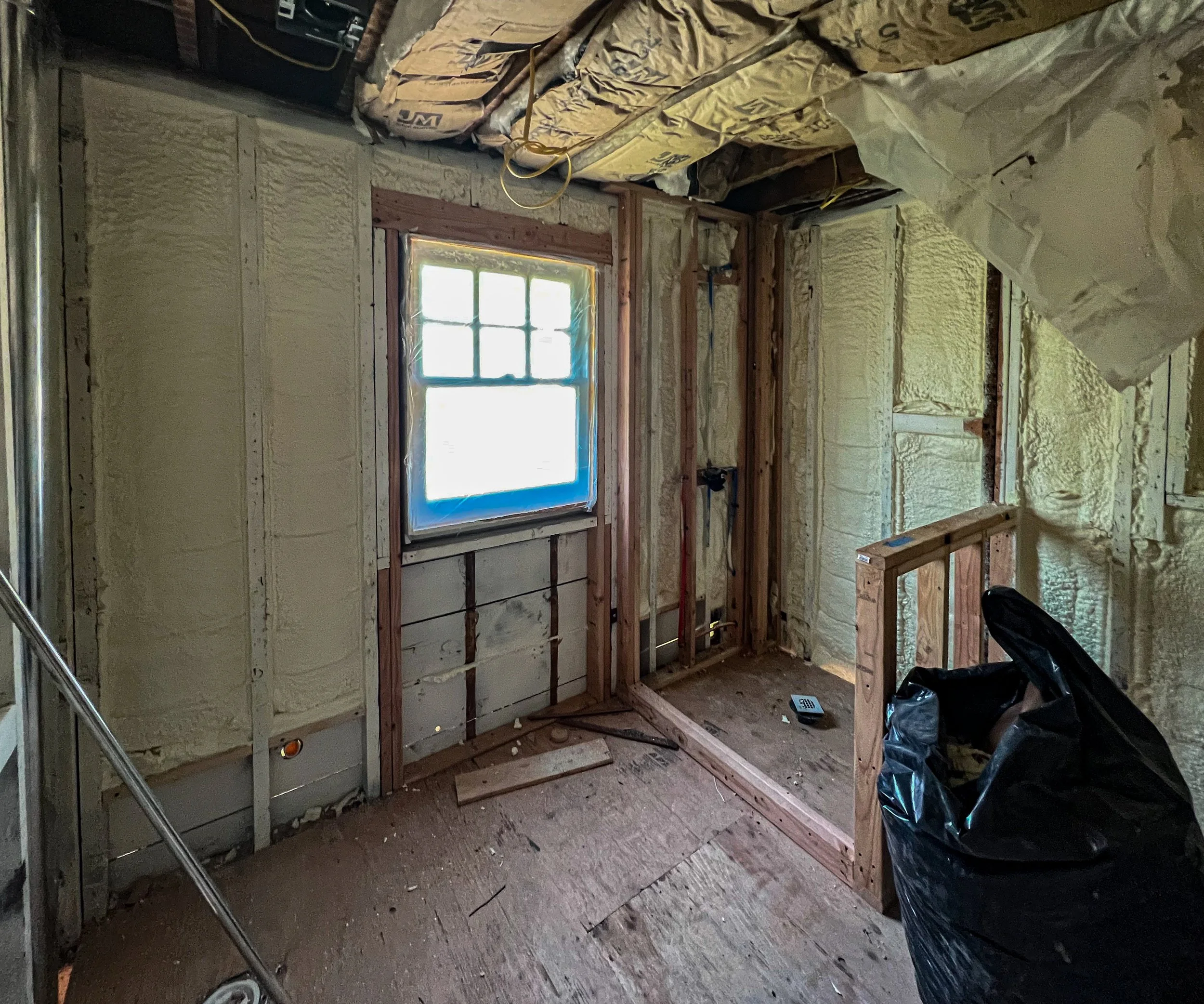 Room under construction with insulation on walls, window with sunlight coming through, wooden framing, construction tools, and a black trash bag.
