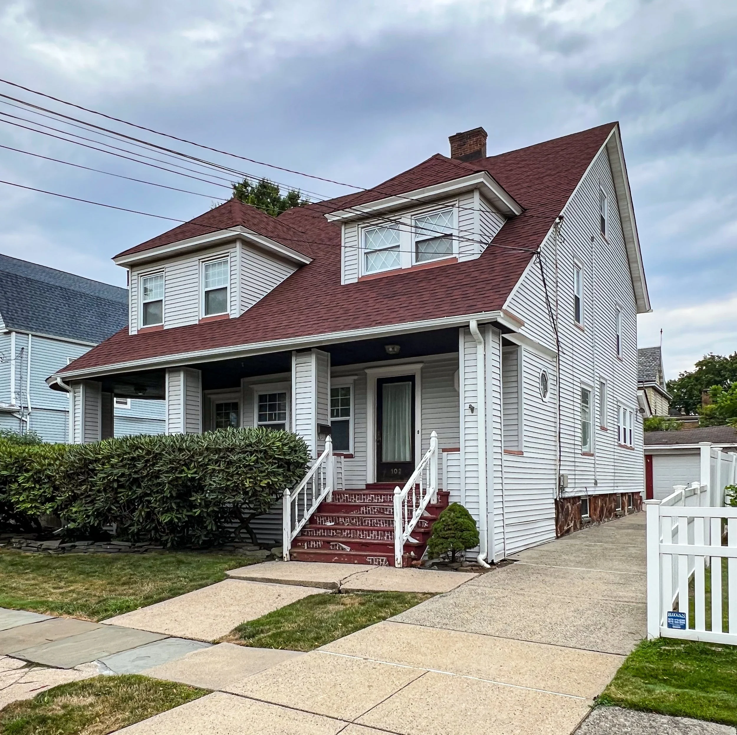 A two-story house with white siding and a red roof, front porch with stairs and bushes, driveway to the right, and overcast sky overhead.