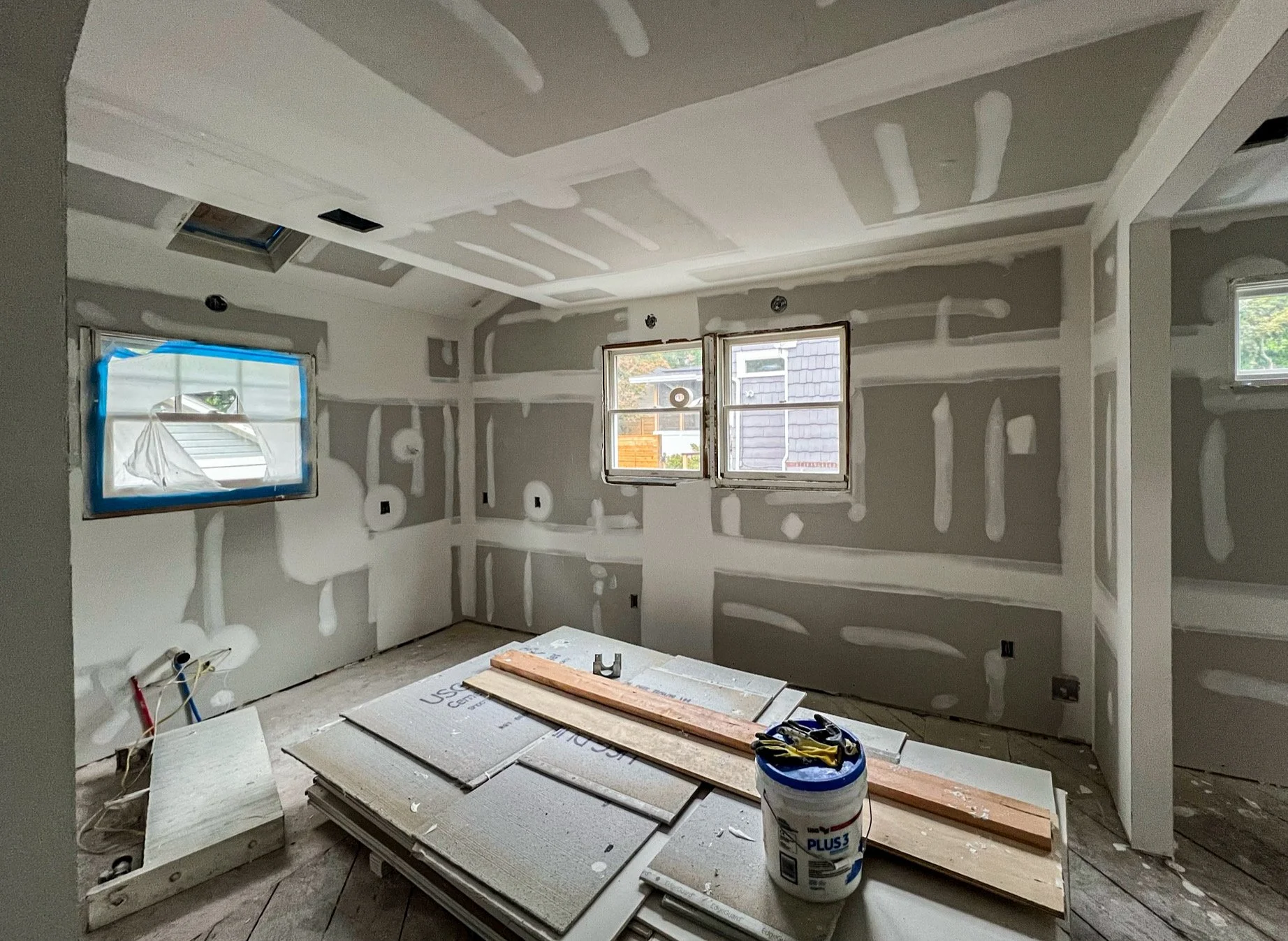 Interior of a room under construction with drywall on the walls and ceiling, windows with protective covering, and construction materials on the floor.