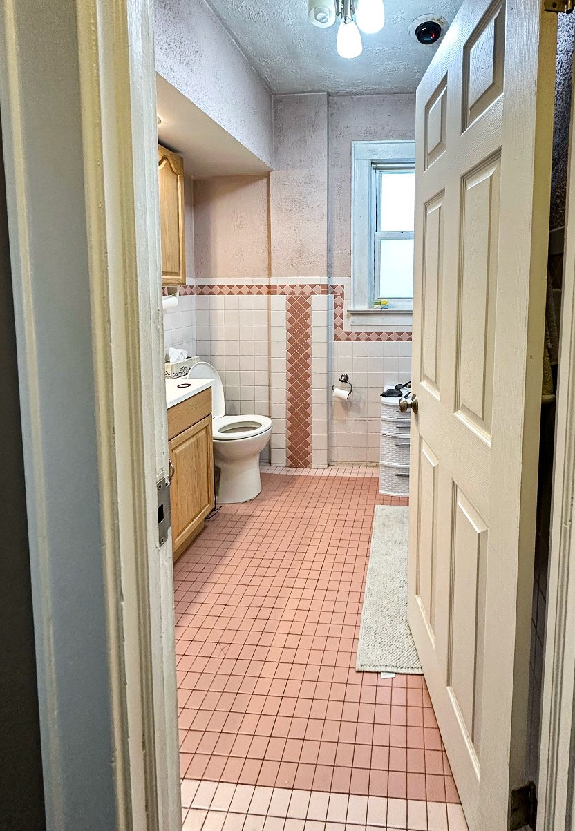 Interior view of a small bathroom taken from doorway, featuring pink tile floor, white and pink wall tiles, a window, toilet, small cabinet, and laundry basket.