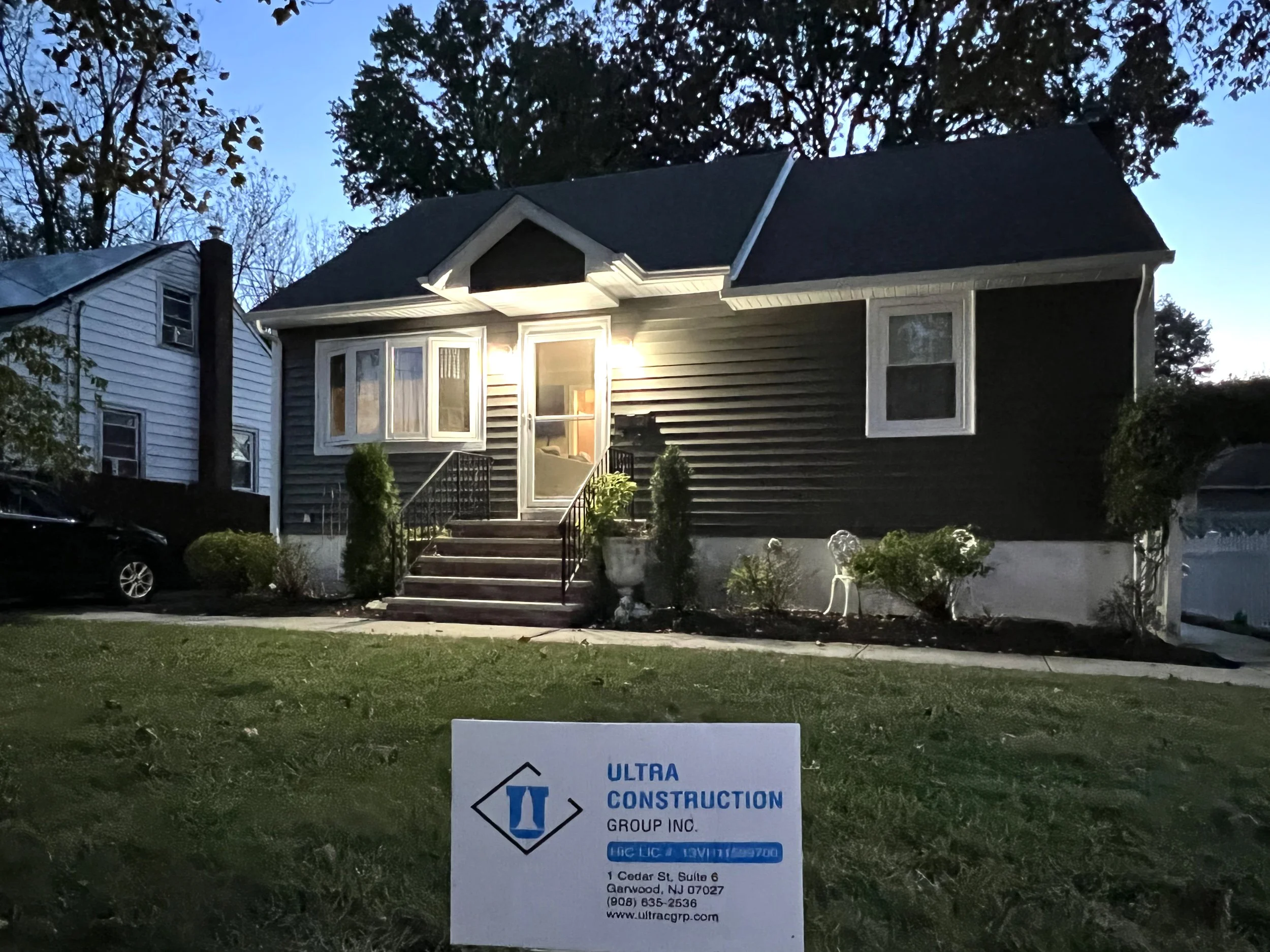A house with dark exterior siding and white trim, illuminated by exterior lights, with a front porch accessed by stairs. The house is surrounded by landscaped bushes and plants. A sign in front of the lawn reads 'ULTRA CONSTRUCTION GROUP INC.' with c