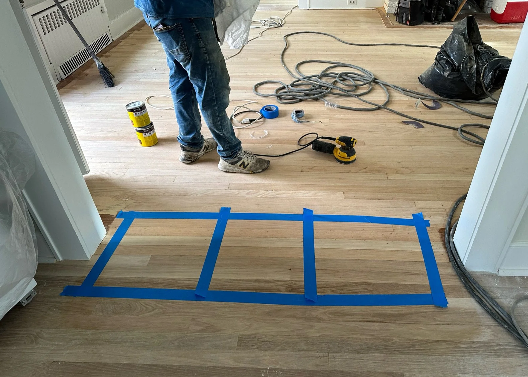 A person standing on a wooden floor with blue painter's tape outlining a rectangular area, surrounded by tools and construction supplies, in a room under renovation.