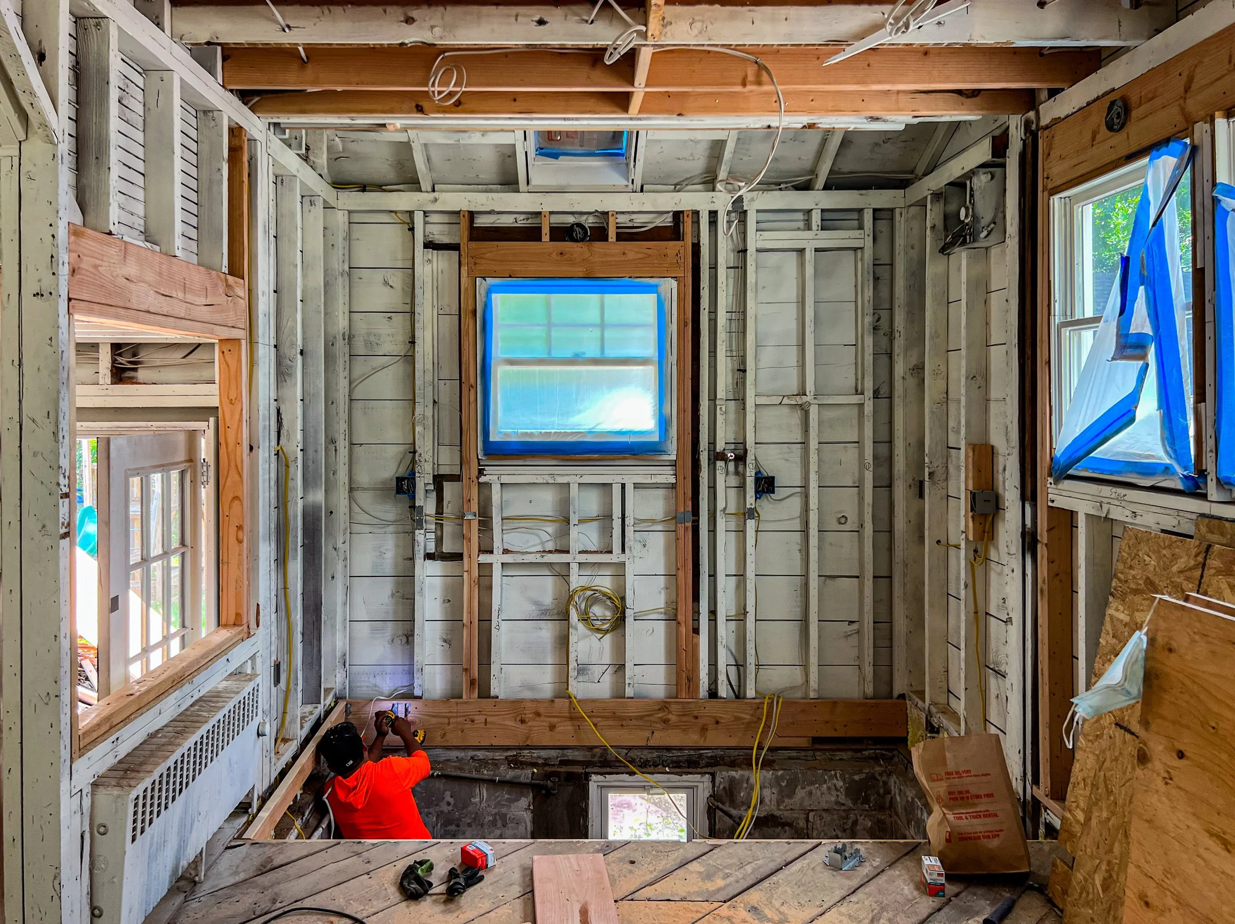 Interior of a house under construction with exposed framing, multiple windows, a worker in an orange shirt working on the wall, and construction tools and materials scattered around.
