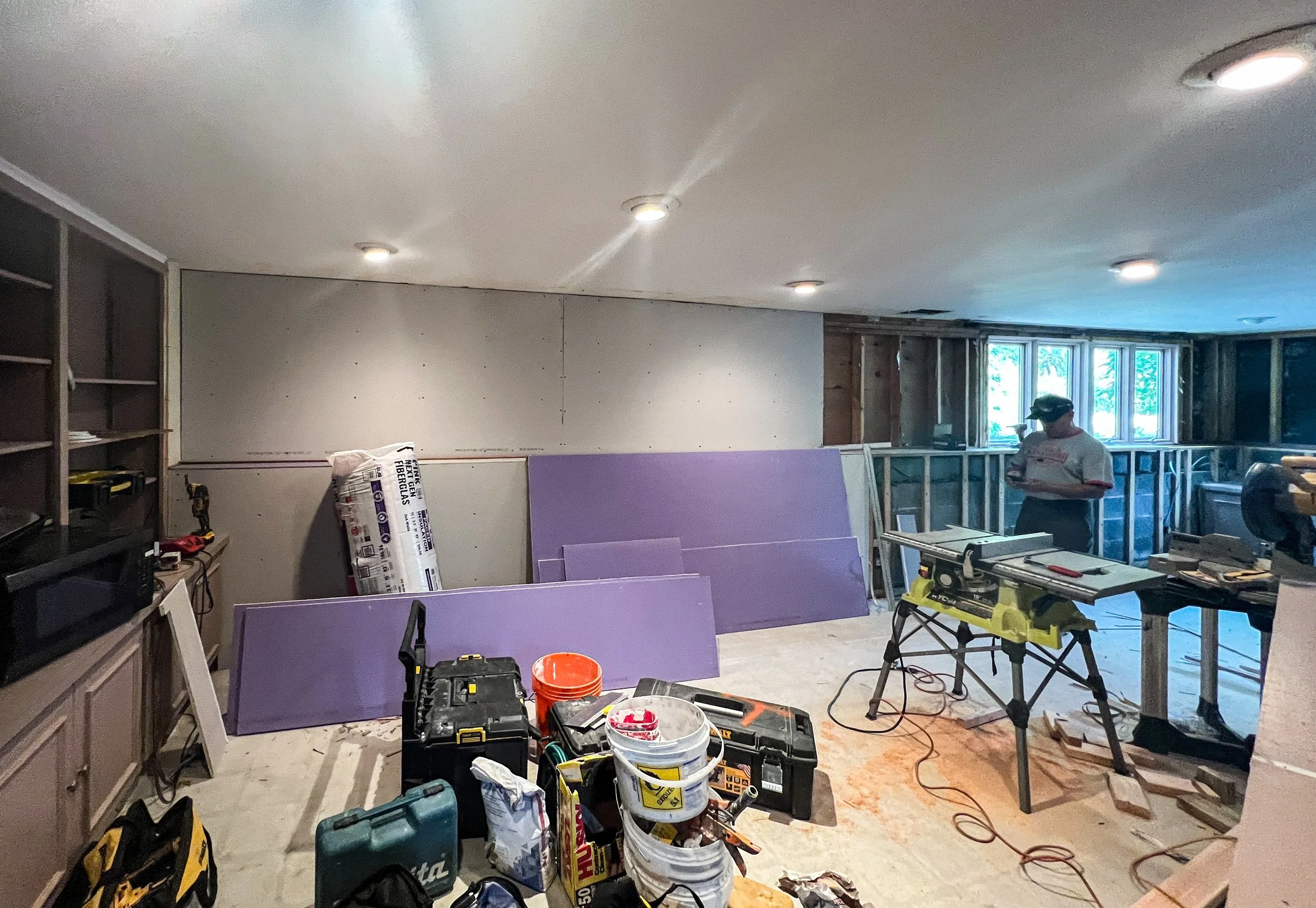 A room under renovation with construction tools, materials, and equipment scattered on the floor. A worker is standing near the windows, which have no wall covering.