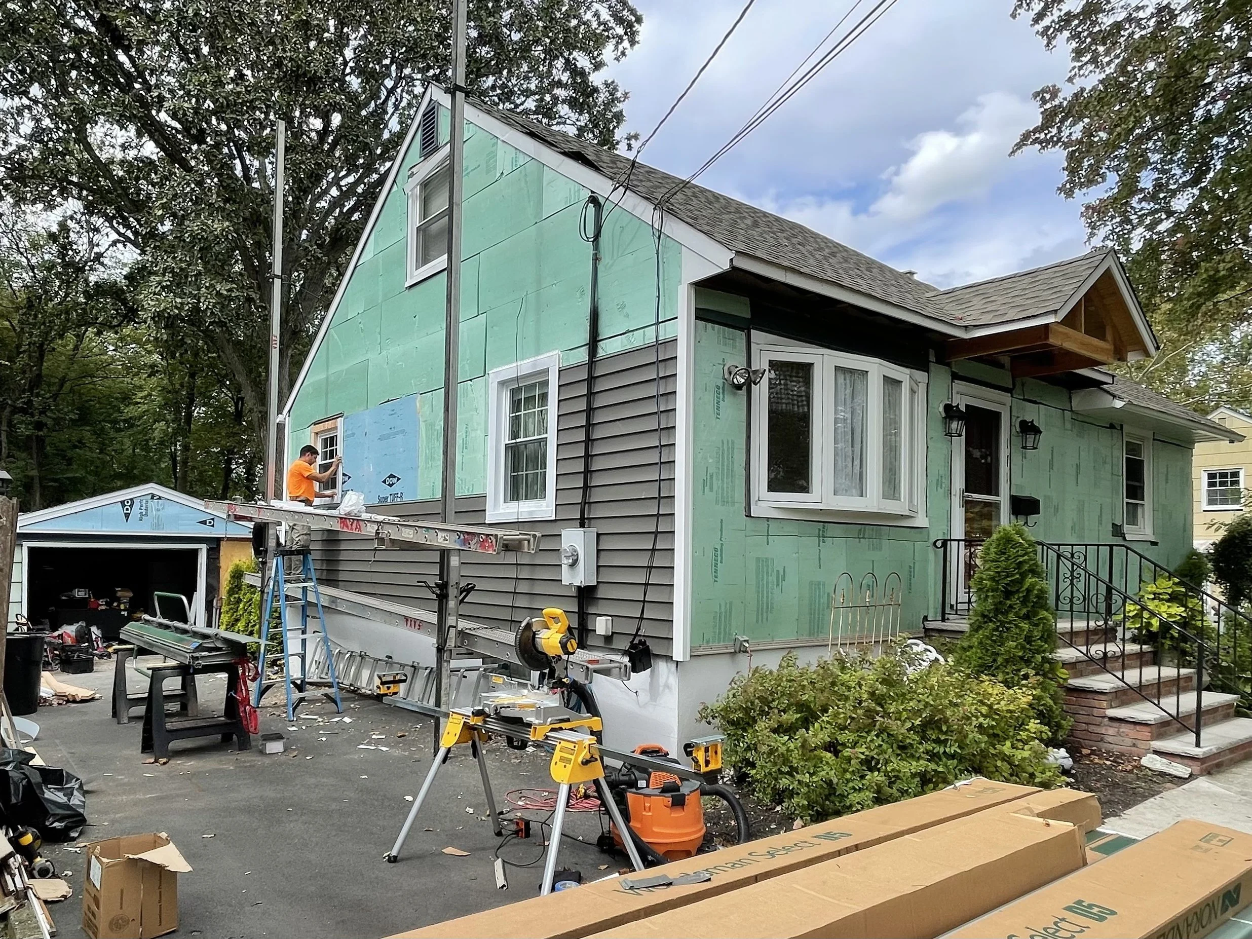 House under renovation with siding being installed, construction tools, and worker on ladder.