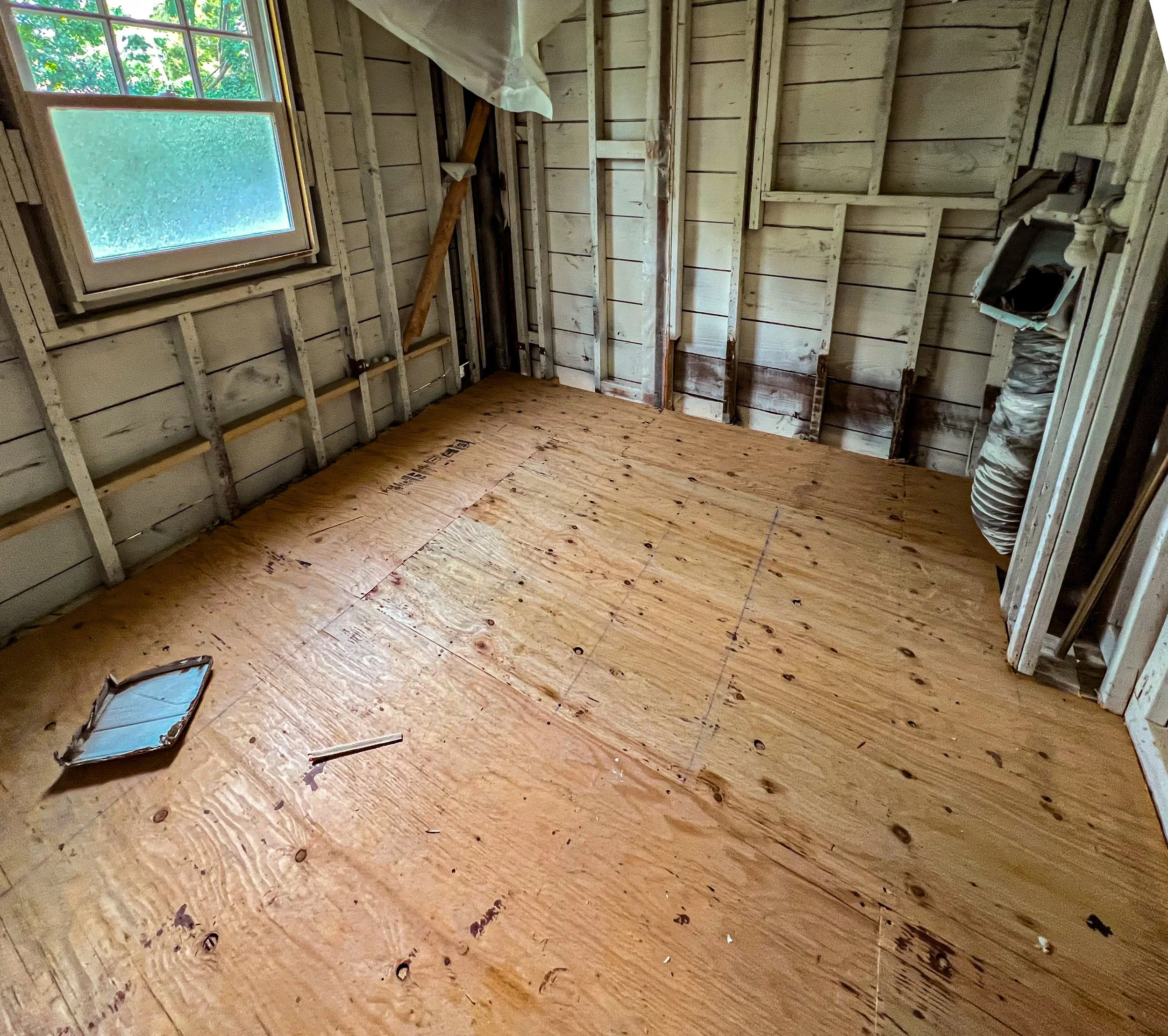 Room under construction with plywood flooring and unfinished wall framing, a window, and construction materials.