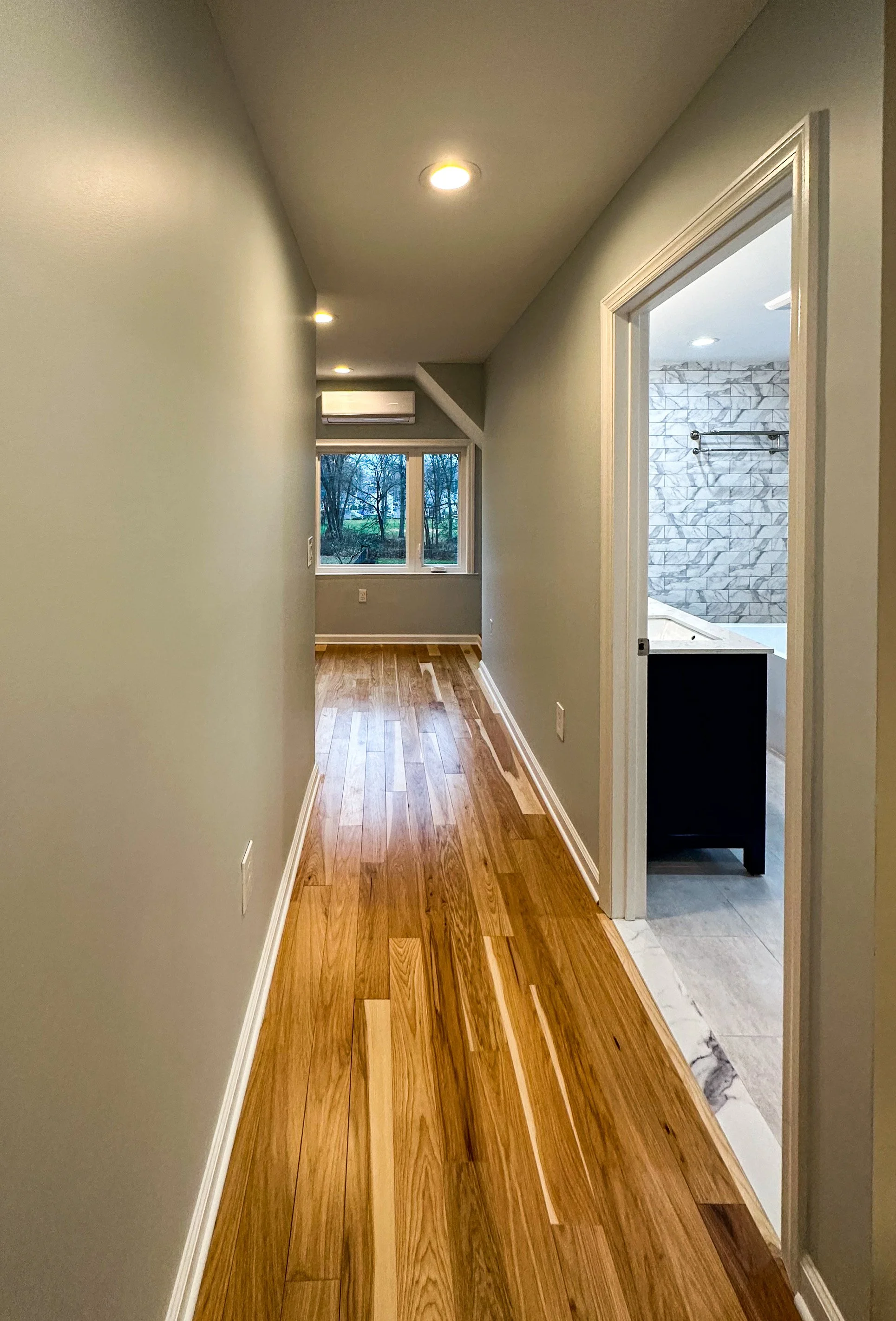 Interior view of a hallway with hardwood floors, gray walls, ceiling lights, and a window at the end, with a bathroom visible on the right side.