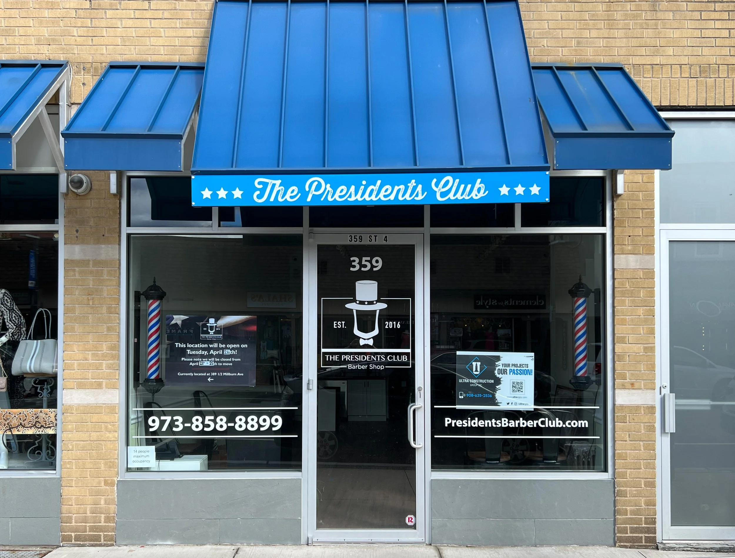 Front view of a barber shop named The Presidents Club with a blue awning, glass door, and various signs on the windows.