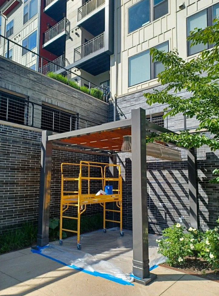 A small outdoor construction or renovation site with a yellow scaffolding, a protective canopy, and a blue bottle of cleaning solution, located against a dark brick wall, with a contemporary apartment building in the background.