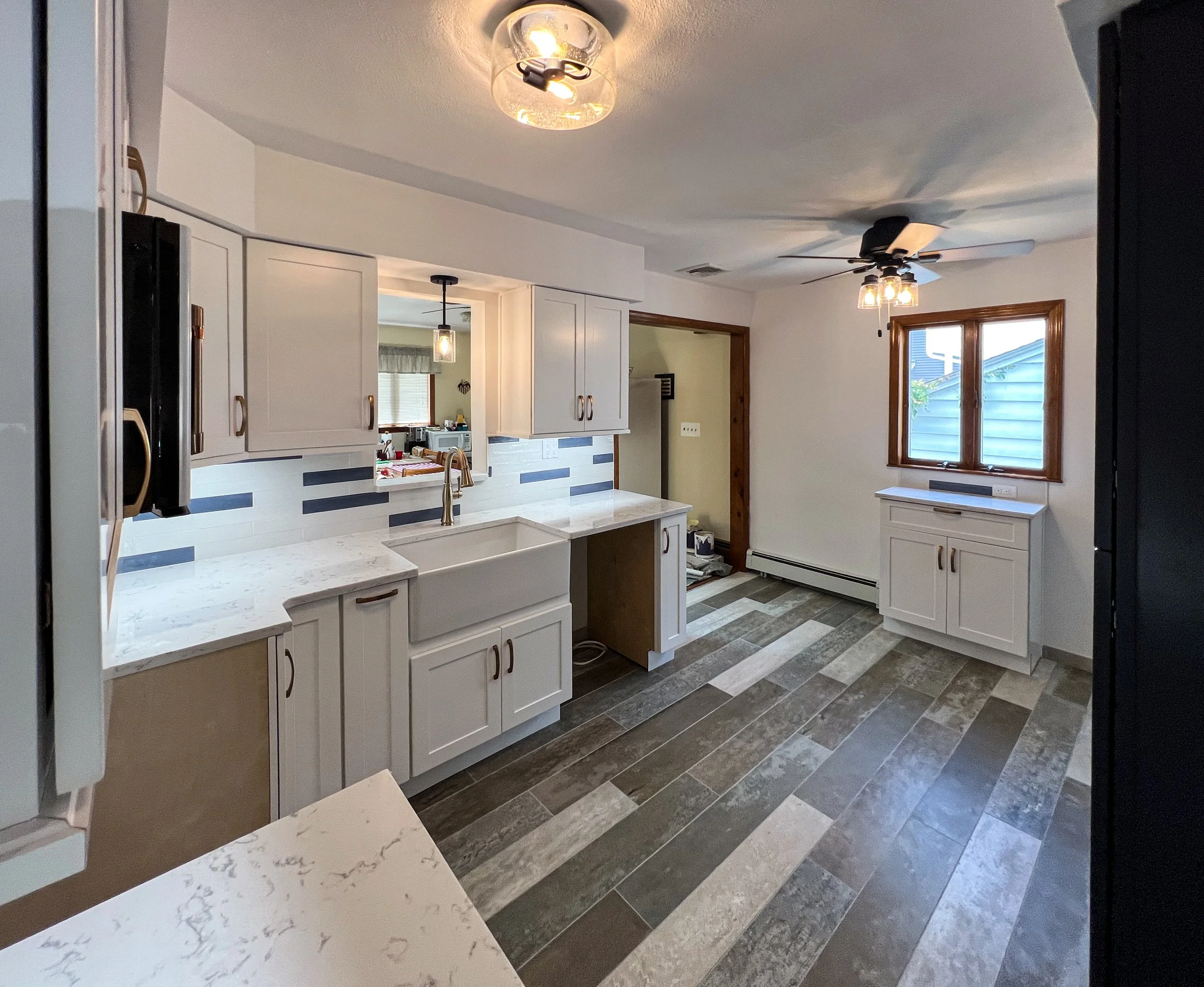 Empty kitchen with white cabinets, marble countertops, a large farmhouse sink, window with wooden trim, and multicolored tiled floor.