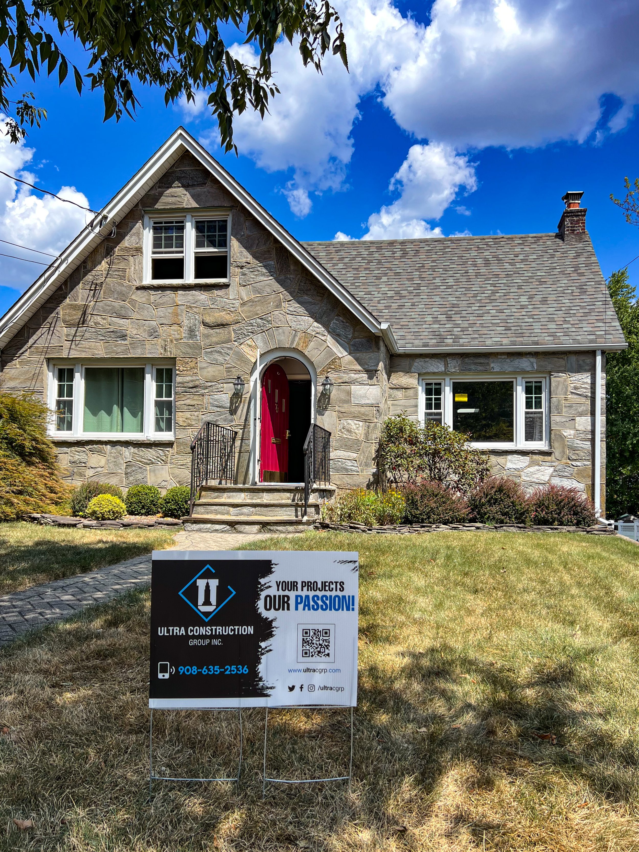 Stone house with red front door, front steps, and a sign in the lawn advertising Ultra Construction Group Inc., with contact information and social media icons, under a partly cloudy blue sky.