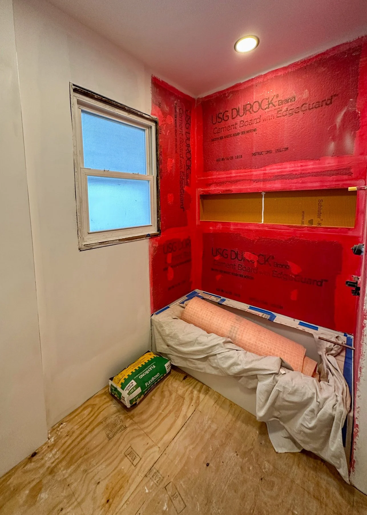 A small room under construction with a window, red drywall, and a cutout shelf. There is a small mattress with a pillow, bedding, and a bag of construction supplies on the unfinished wooden floor.