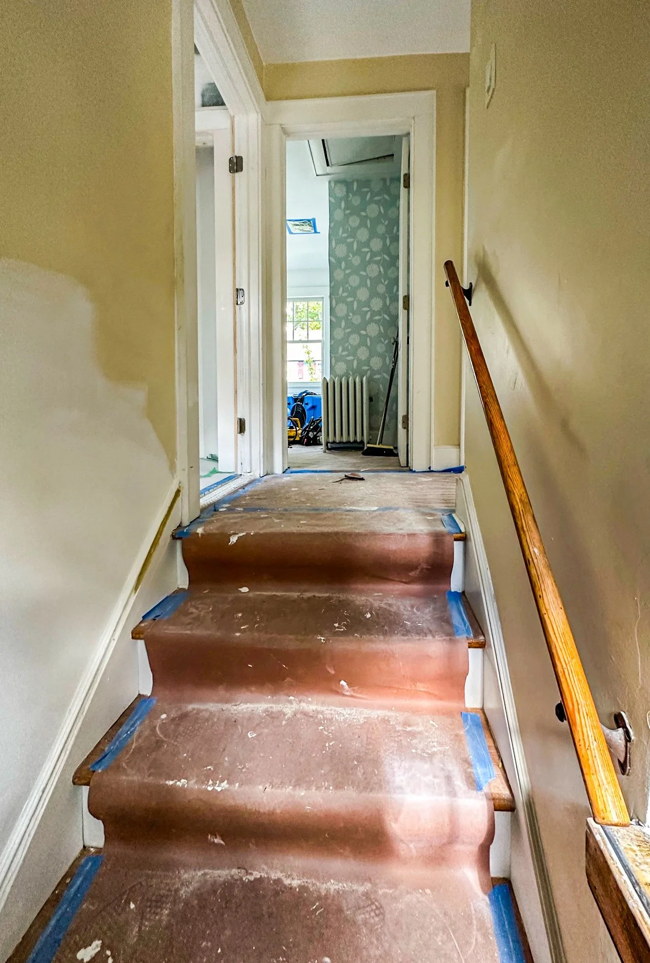 Interior of a house under renovation showing a stairway with brown protective covering, a wooden handrail on the right, and open doorways leading to a sunlit room in the background.