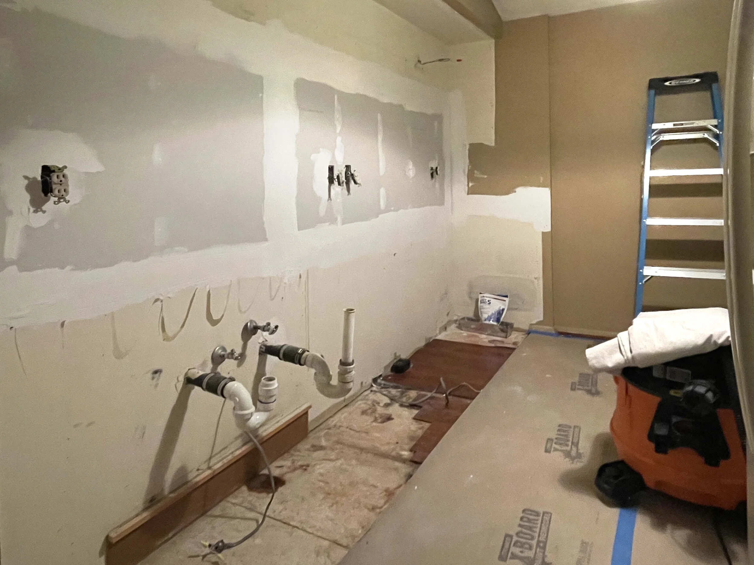 A kitchen renovation in progress. The wall has drywall with visible drywall joints and cutouts for appliances or outlets. Plumbing pipes are exposed below the wall. Construction tools and materials, including a ladder, a tool bag, and a bag of joint 