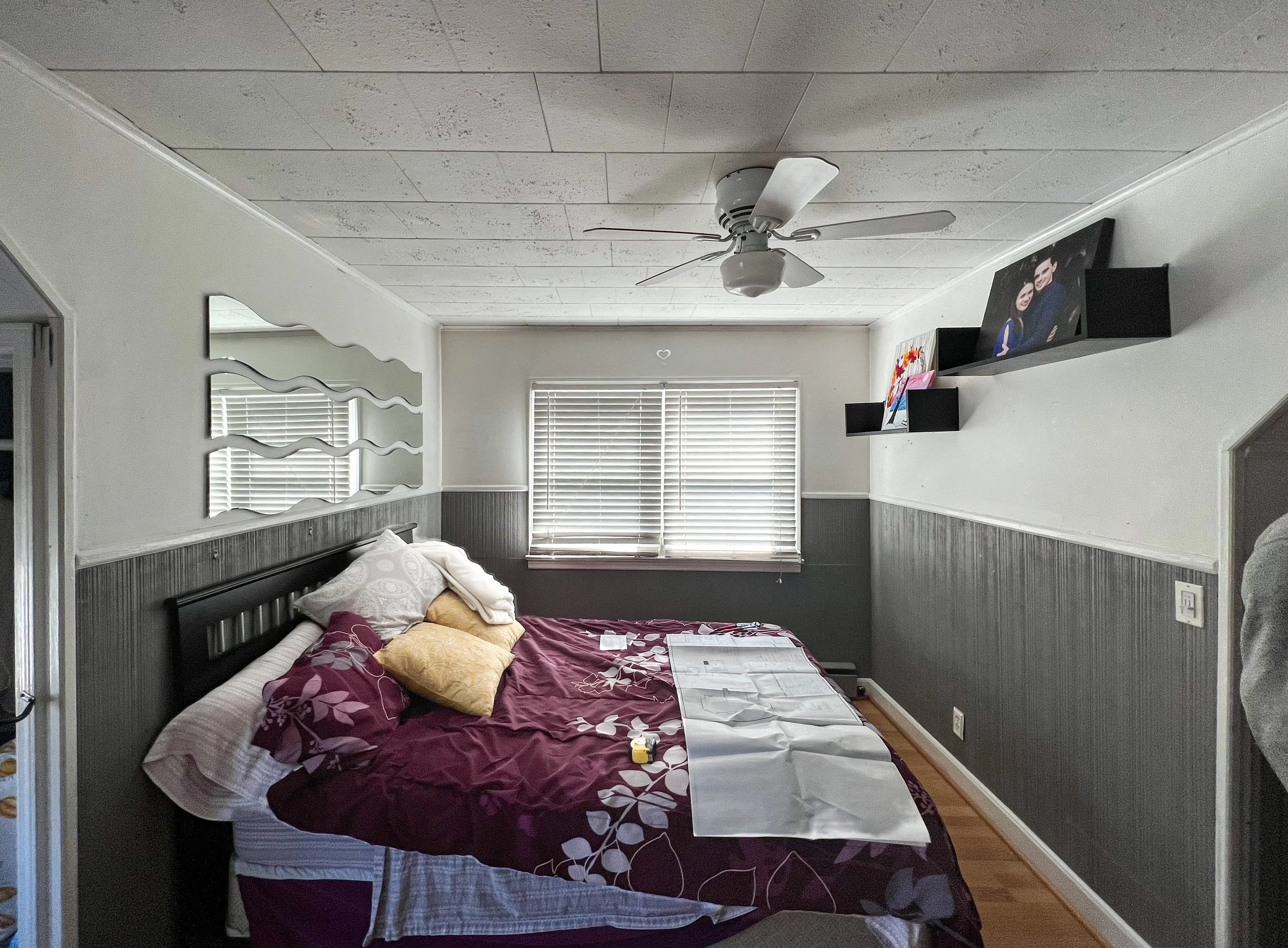 A bedroom with a bed covered in a maroon and white floral comforter, several pillows, a window with blinds, a ceiling fan, and wall-mounted shelves with photo frames and artwork.