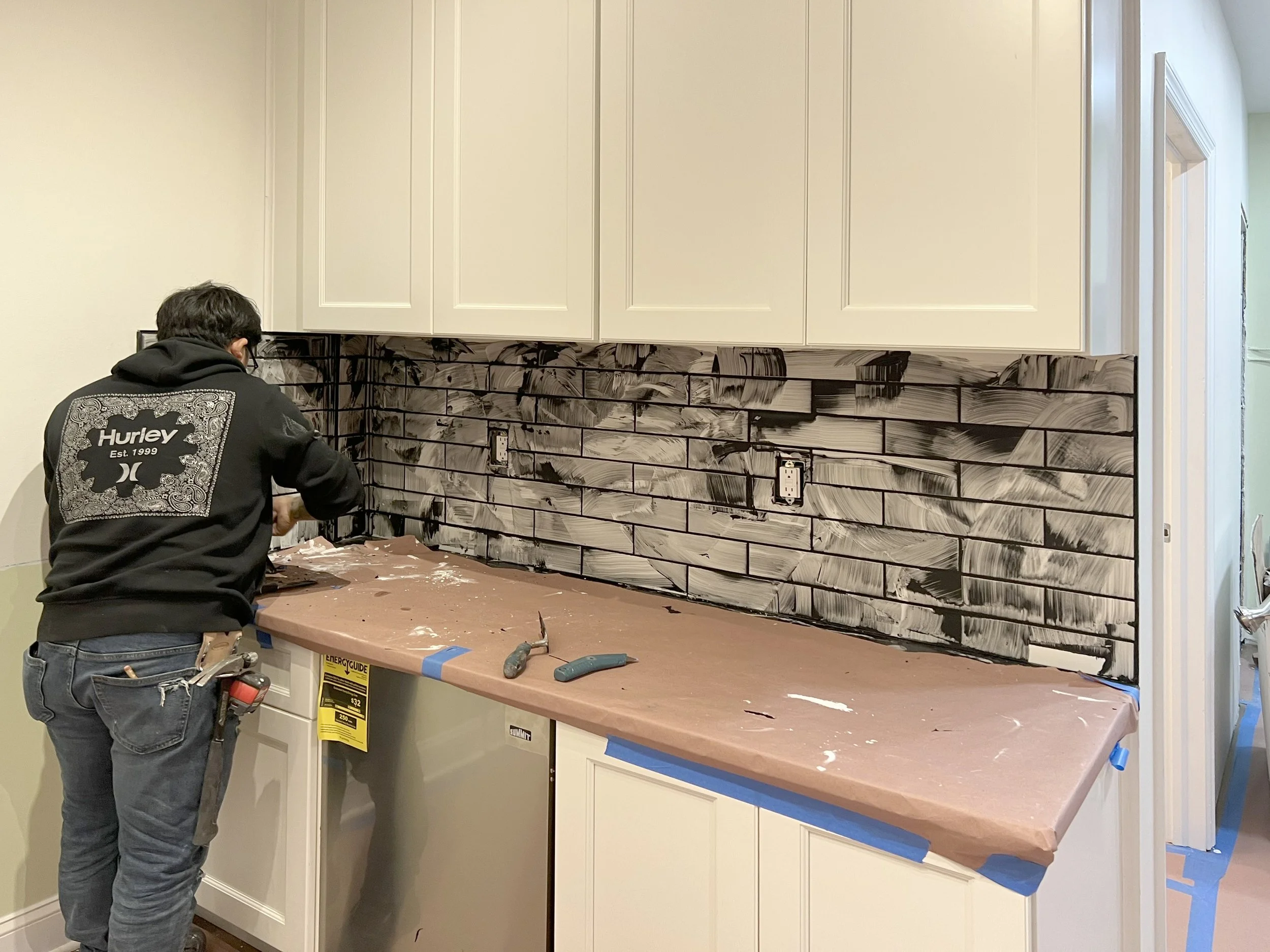 A person working on a kitchen backsplash with black and white painted brick pattern, tools and tape on the countertop, and white upper cabinets.
