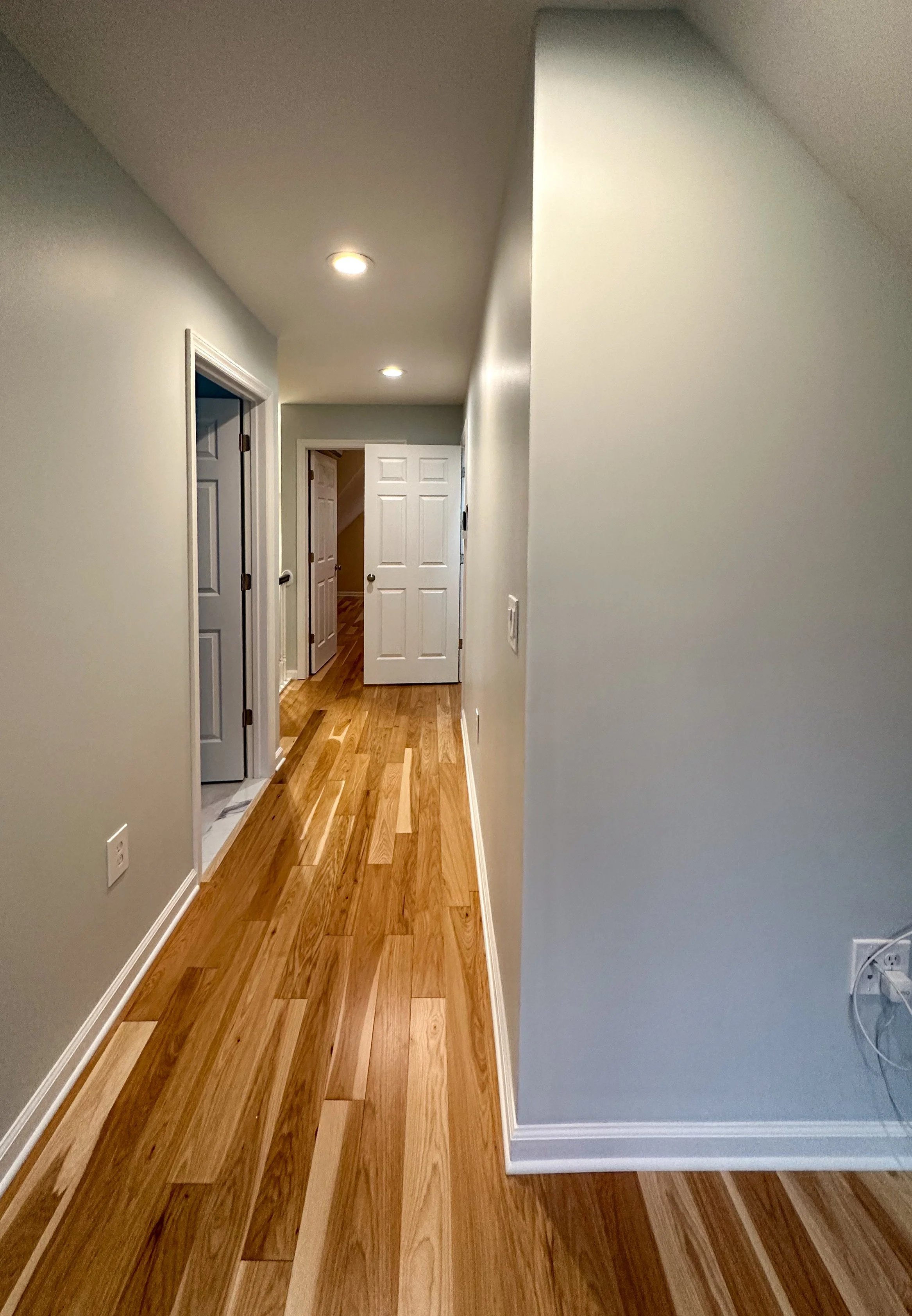 A hallway with hardwood floors, white baseboards, light-colored walls, several closed doors, and ceiling lights.