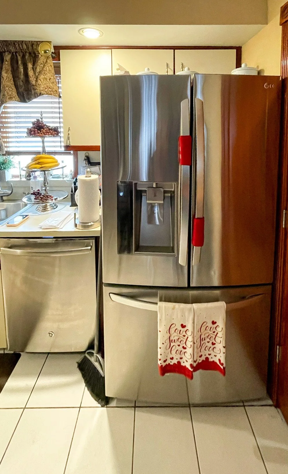 Stainless steel refrigerator with a towel hanging on the handle that says 'love sweet love'. In the background, a kitchen with a window, curtains, a fruit bowl with bananas and grapes, paper towels, and various kitchen appliances.