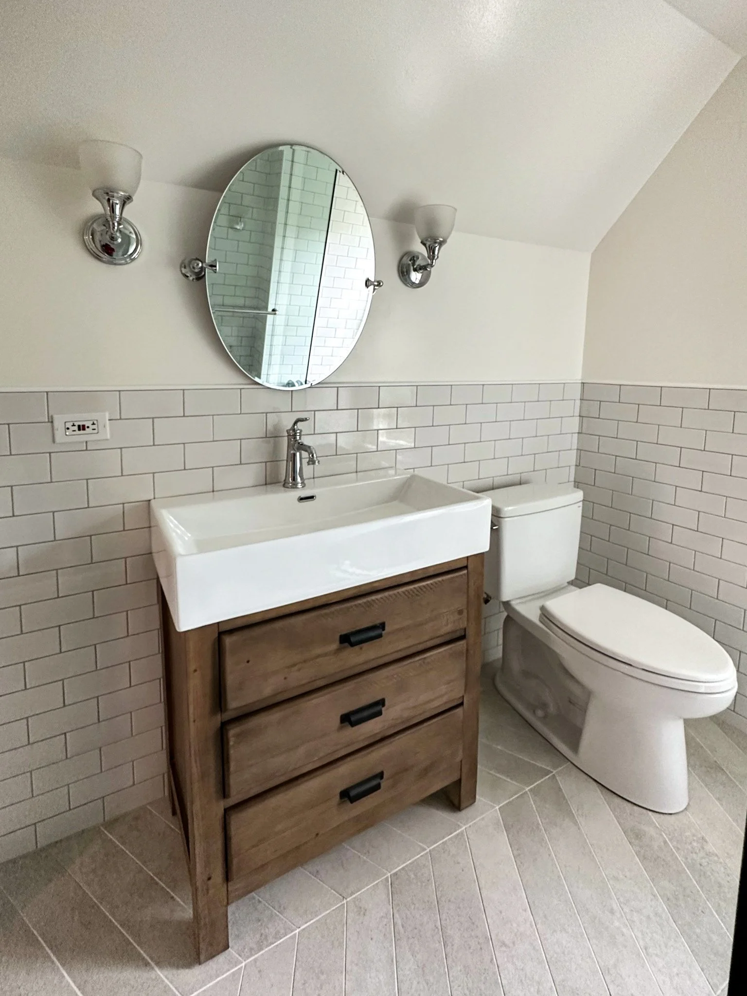 Bathroom with white ceramic sink vanity, wooden drawers, oval mirror, white toilet, white tiled half-wall, wall-mounted light fixtures, and an electrical outlet.
