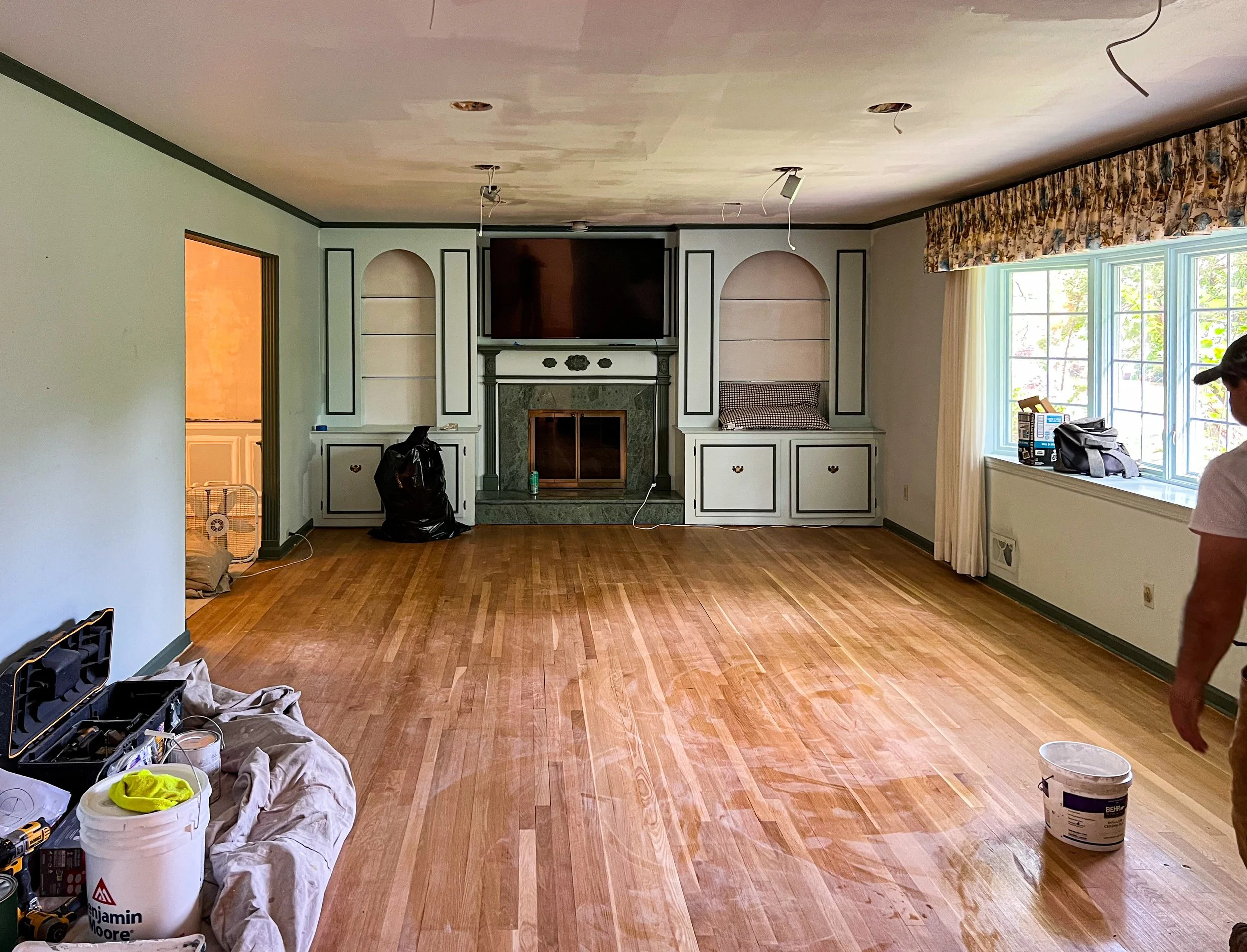 Living room under renovation with hardwood floor, built-in cabinets around a fireplace, and a large window with floral curtains. Construction tools and supplies are scattered around.