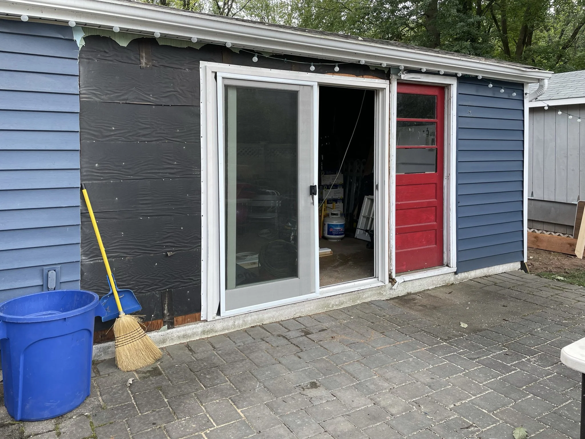 Side of a house with a blue exterior, black insulation material, a sliding glass door, and a red wooden door. A yellow broom with a natural straw head is leaning against a blue bucket, placed on a brick patio.