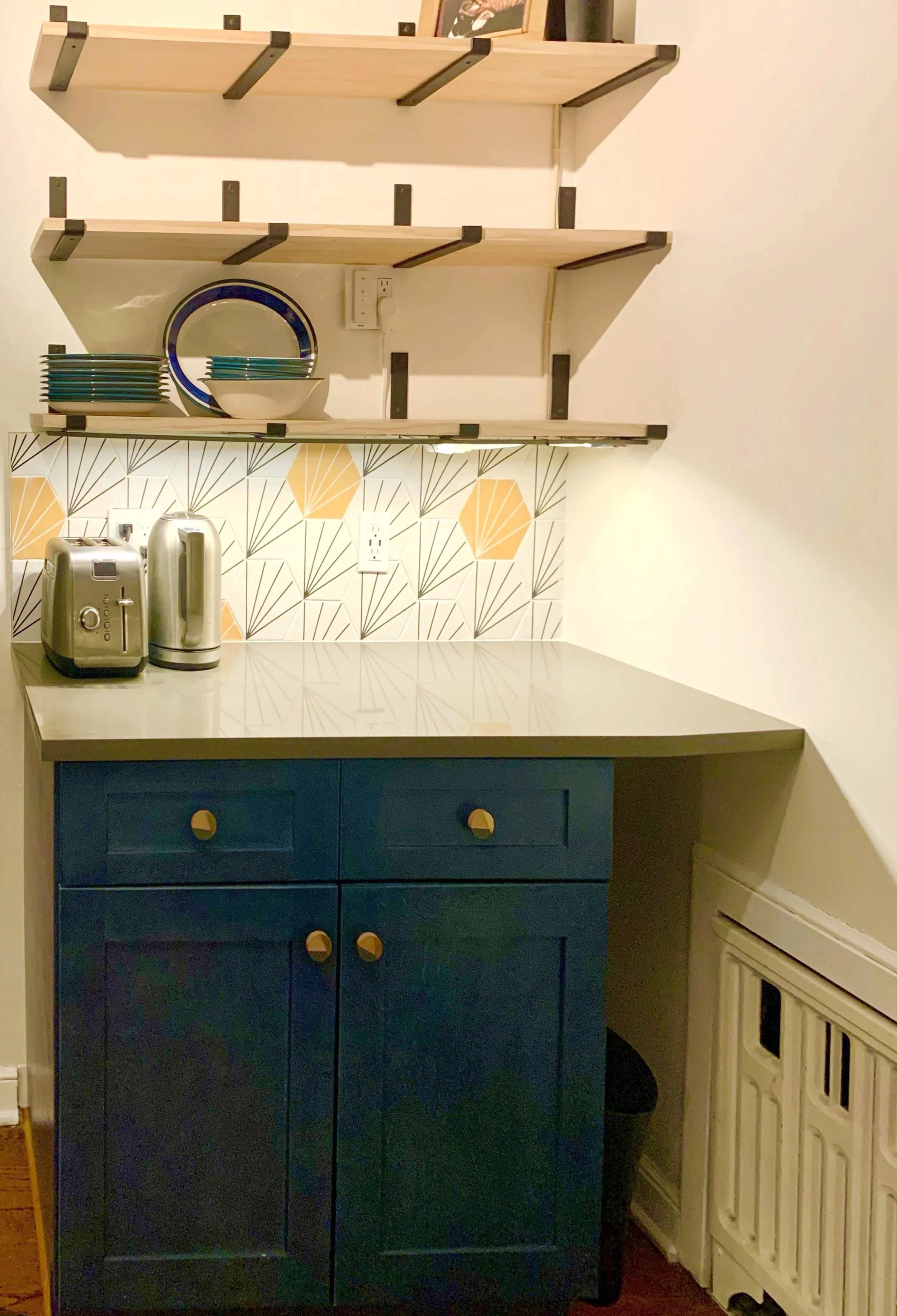 Kitchen corner with navy blue cabinet, stainless steel toaster and electric kettle, patterned tile backsplash, two wooden shelves with plates and bowls, and a framed photograph on the top shelf.