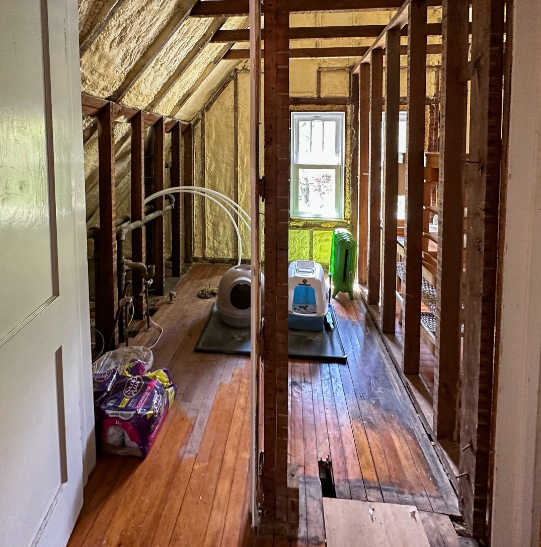 A room under renovation with exposed wooden framing, floorboards, a window, and a sloped ceiling with insulation. There are pet supplies and a stack of green plastic chairs on the floor.