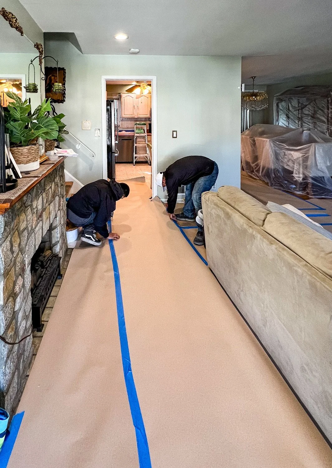 Two people are measuring the floor of a living room with blue painter's tape. A couch is on the right side, and a stone fireplace is on the left. There is a doorway leading to a kitchen in the background.